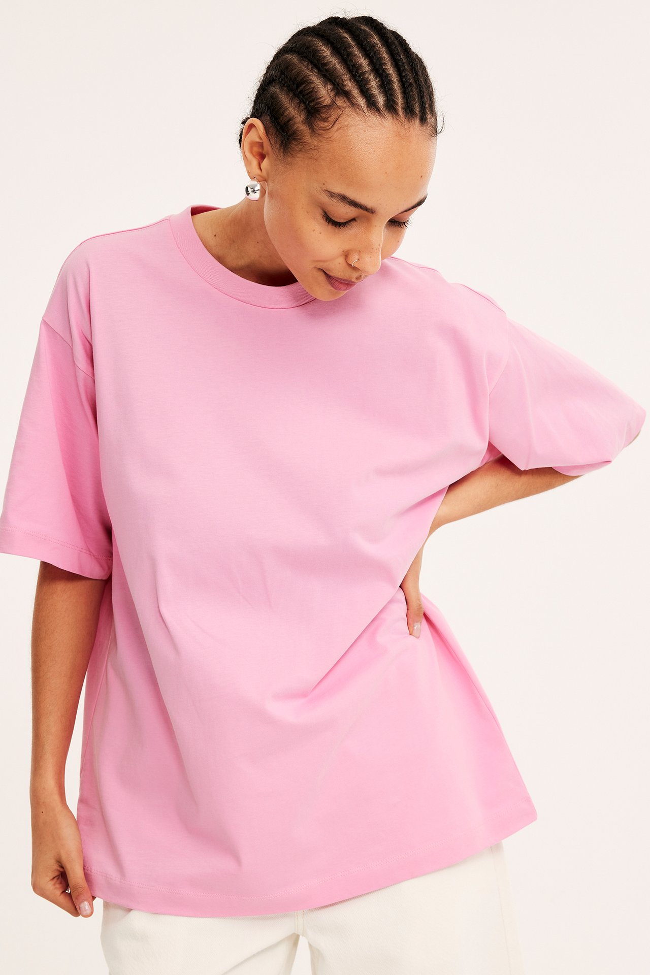 Framifrån, kvinna i en ljusrosa oversized t-shirt i bomull, ljusa byxor och silverörhängen.