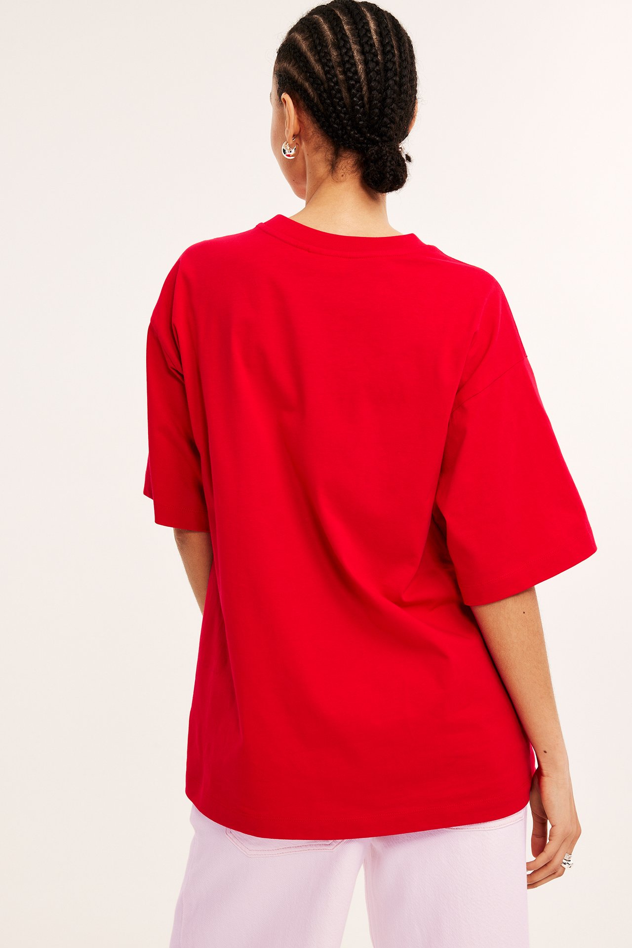 Bakifrån: kvinna i röd oversized bomulls-T-shirt, ljusrosa byxor, silverörhängen och en ring.