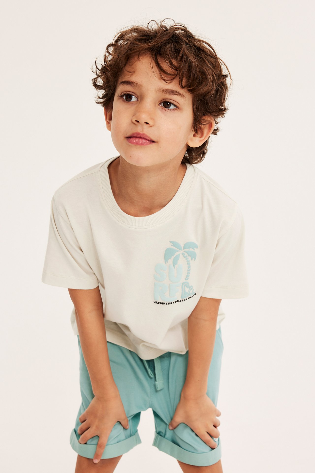 Forfra, et barn i lys beige bomulls-T-skjorte med lyseblått surf-trykk og lyseblå shorts.