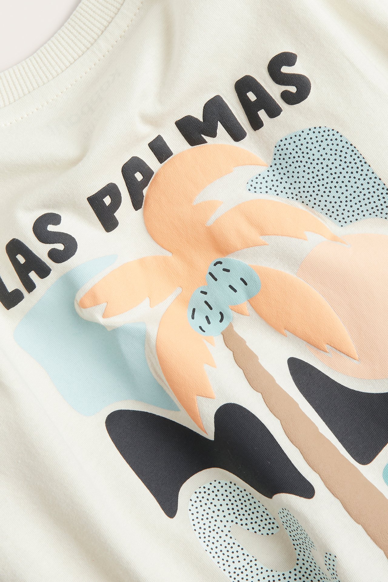 Nærbilde av en lys beige bomulls-t-skjorte for barn med fargerik 'LAS PALMAS'-tekst og palmetrykk.