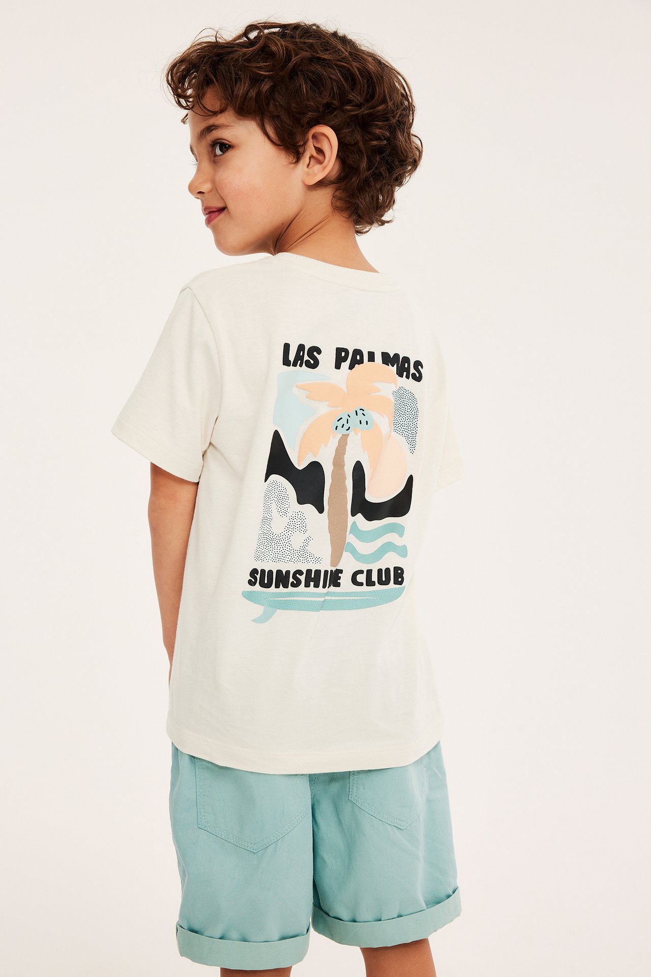 Bakfra: Et barn har på seg en lys beige bomulls-t-skjorte med fargerikt surfprint og lyseblå shorts.
