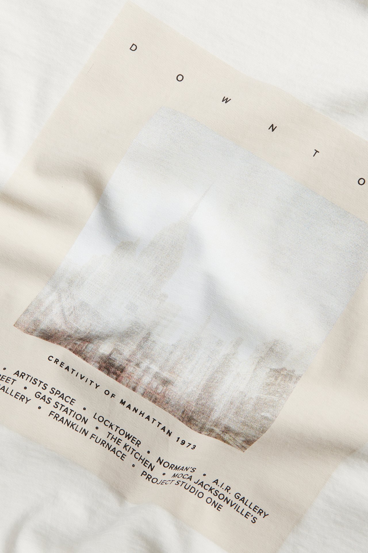 Zbliżenie z przodu na bawełniany T-shirt w kolorze ecru z dużym abstrakcyjnym nadrukiem i czarnym tekstem.