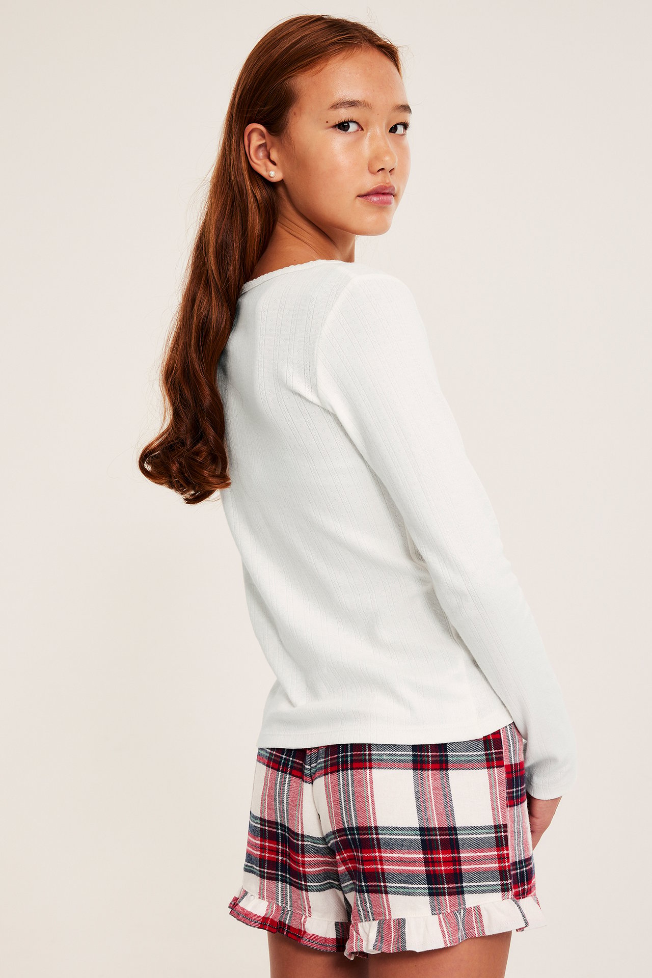 Sidovy av en tjej i offwhite långärmad pointelle pyjamastopp med picotkant och rutiga shorts.