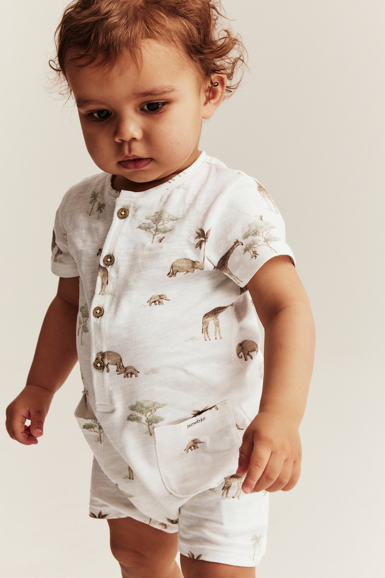 Frontvy av baby i offwhite bomullsromper med safari-djurmönster, knappar framtill och fickor.