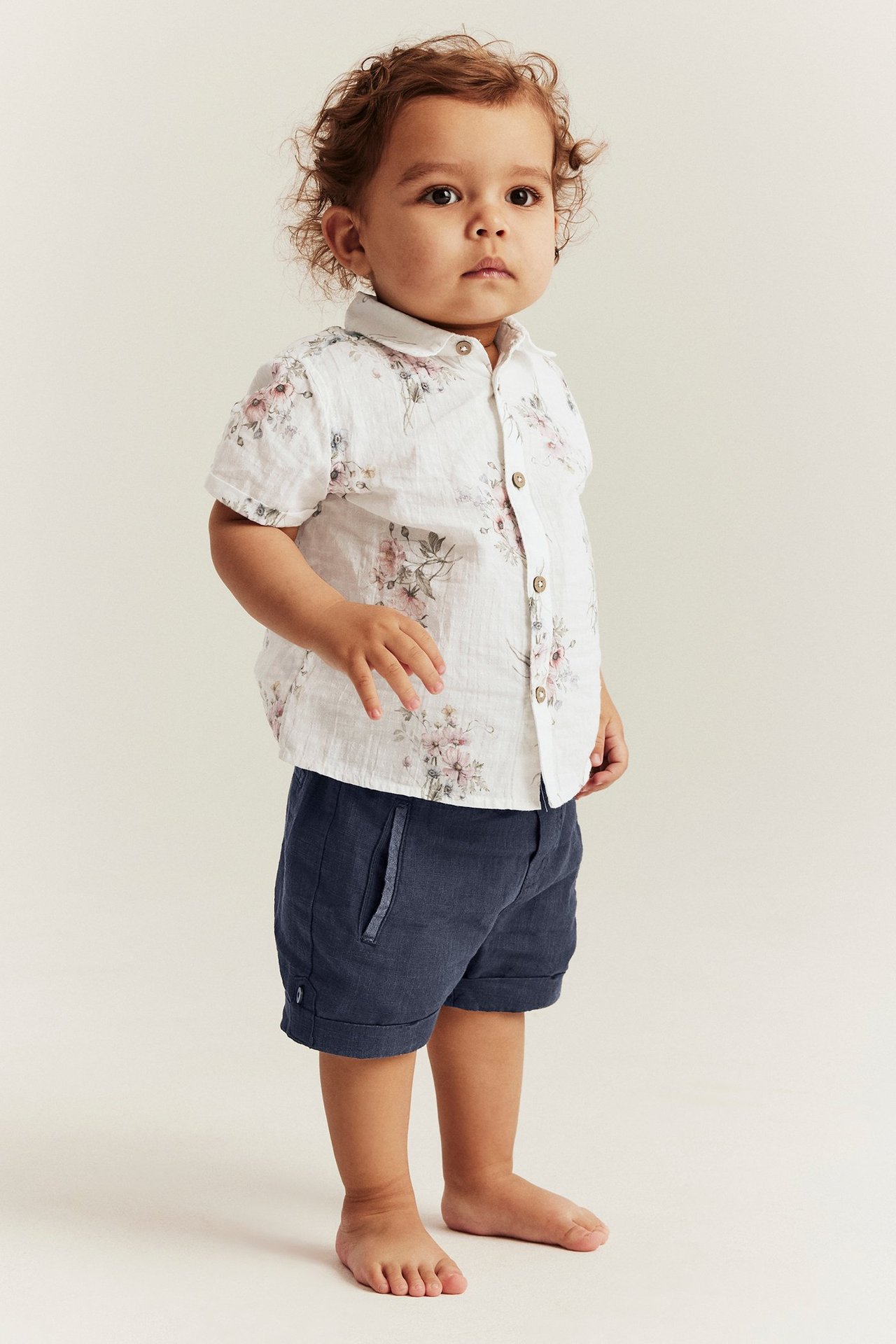 Edestä kuvattu vauva tummansinisissä kudotuissa puuvillashortseissa ja valkoisessa kukkapaidassa.