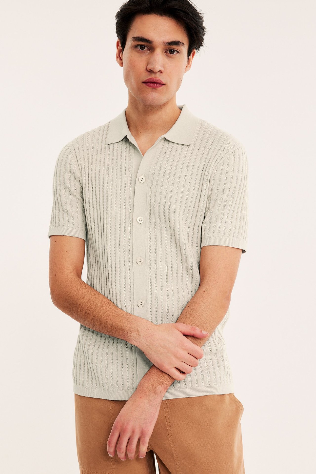 Edestä, mies yllään vaaleanbeige ribbineulottu lyhythihainen napillinen pikeepaita ja ruskeat housut.