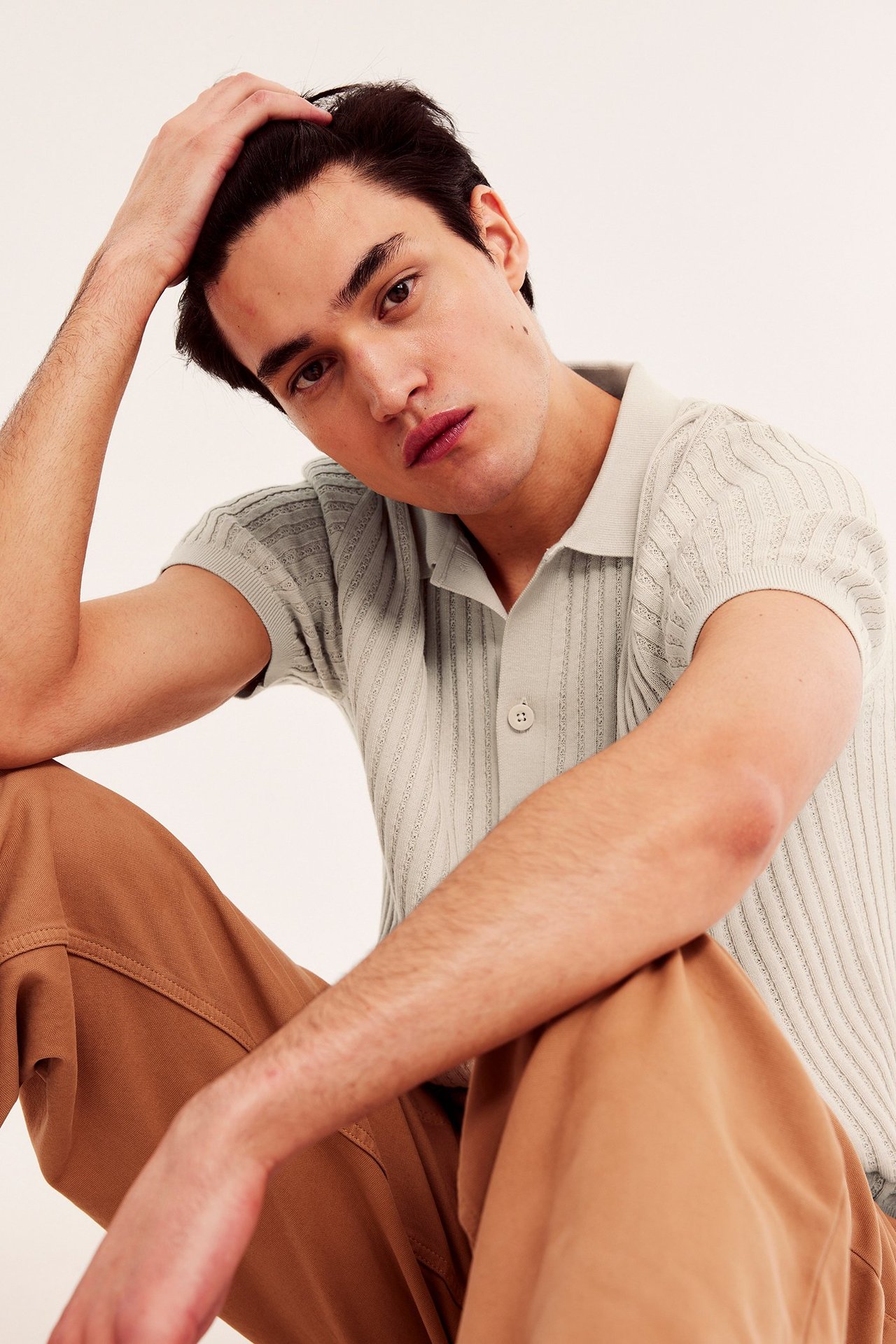 Lähikuva miehestä yllään vaaleanbeige ribattu neulepaita lyhyillä hihoilla ja ruskeat housut.