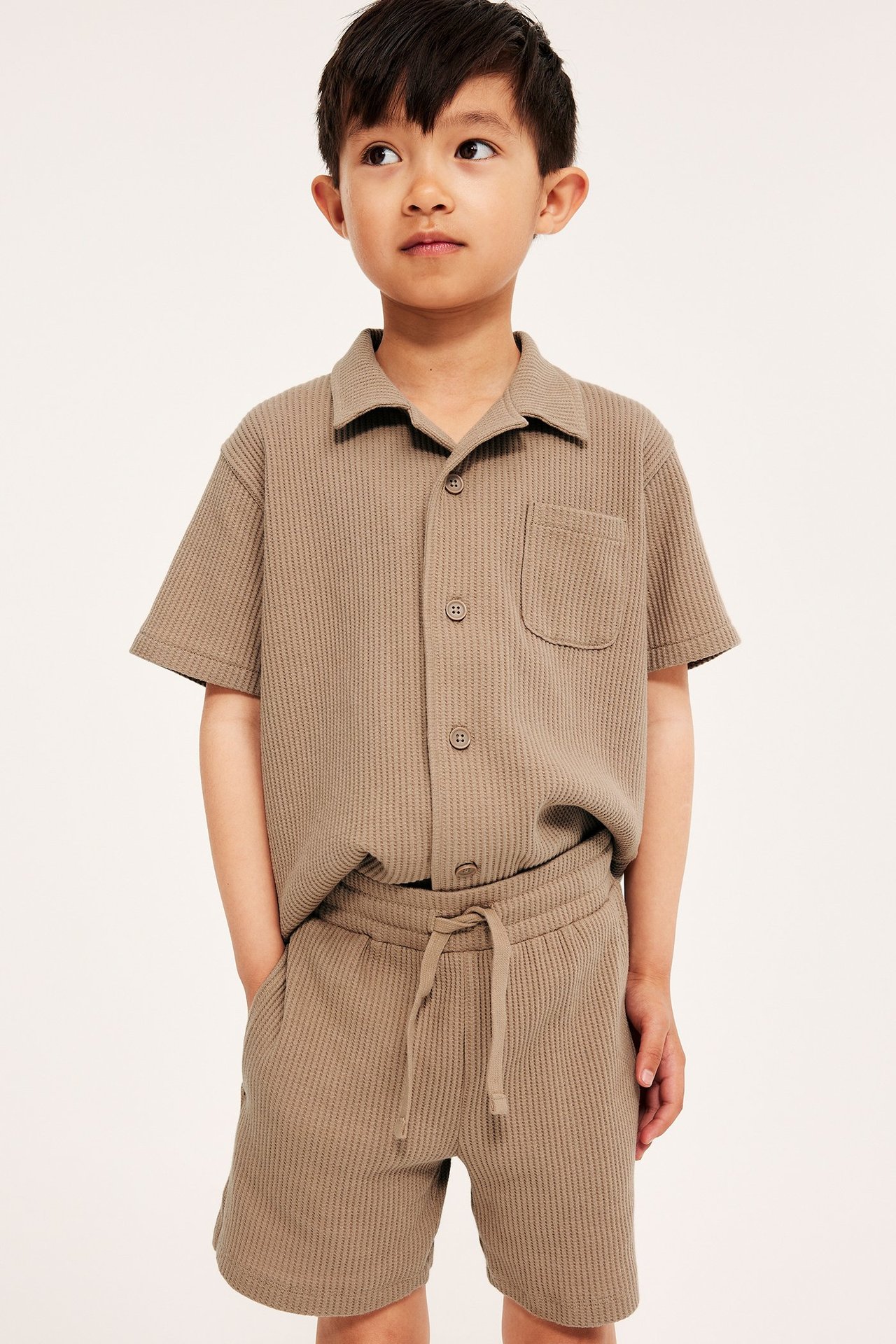 Framifrån: Barn i bruna våfflade bomullsshorts med dragsko, matchande kortärmad skjorta.