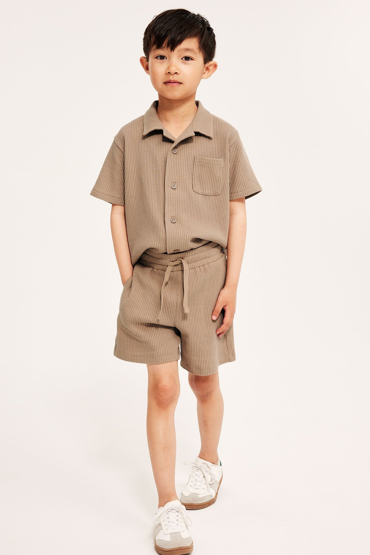 Framifrån: ett barn i bruna våfflade bomullsshorts med dragsko och matchande kortärmad skjorta.