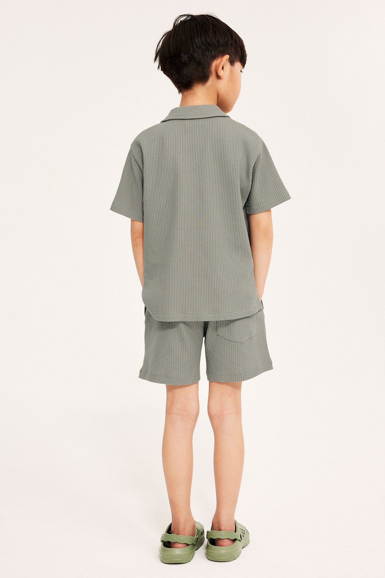 Bakfra av et barn i grønne vaffelshorts, matchende kortermet vaffelskjorte og grønne sandaler.
