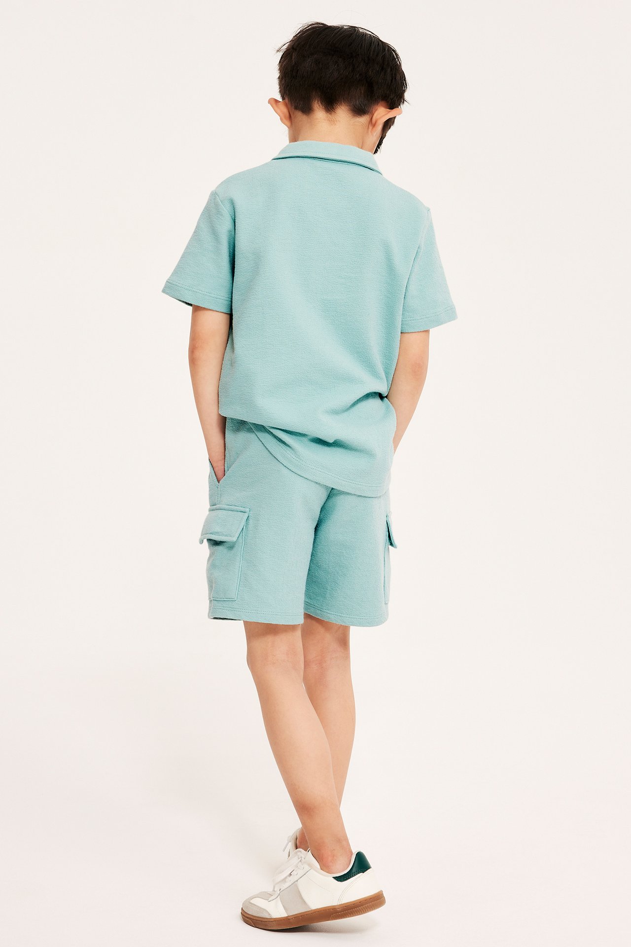 Bakifrån, ett barn i ljusgröna creppade bomullscargoshorts, matchande pikétröja och vita sneakers.