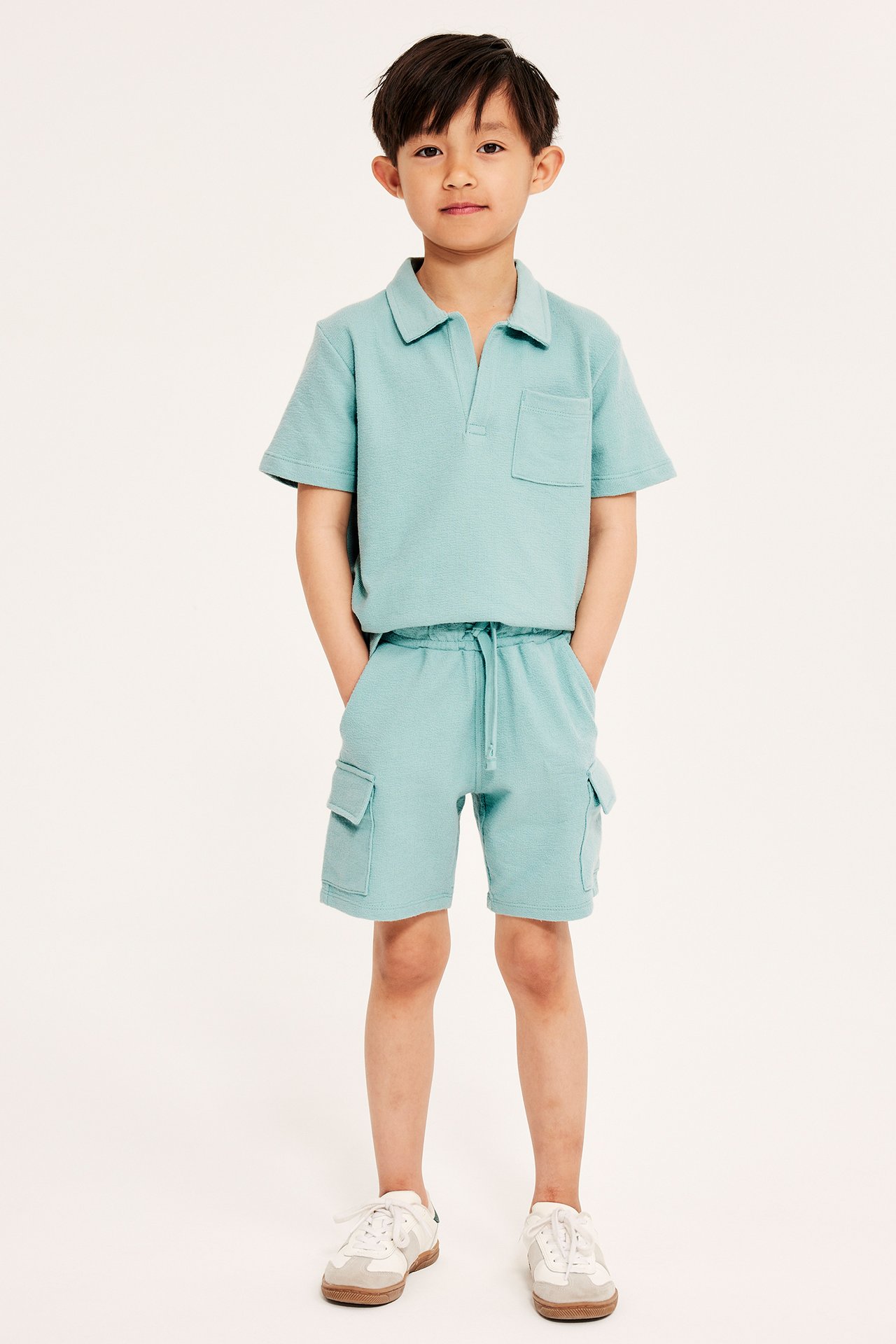 Framifrån: Ett barn i ljusgröna cargoshorts i bomull med dragsko, matchande pikétröja och vita sneakers.
