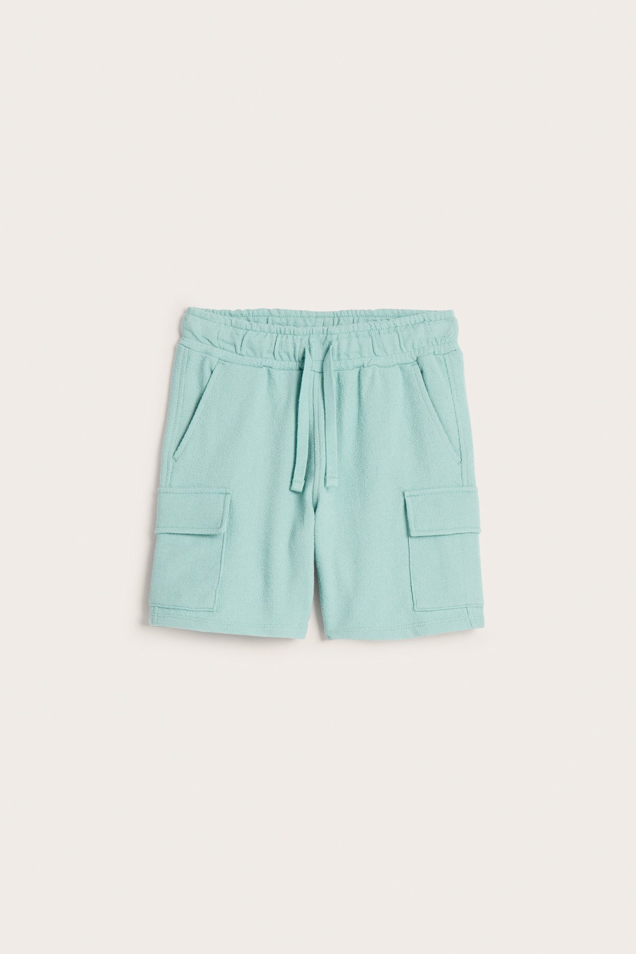Frontvy av ljusgröna mjuka cargoshorts för barn i bomull med resår i midjan, dragsko och fickor.