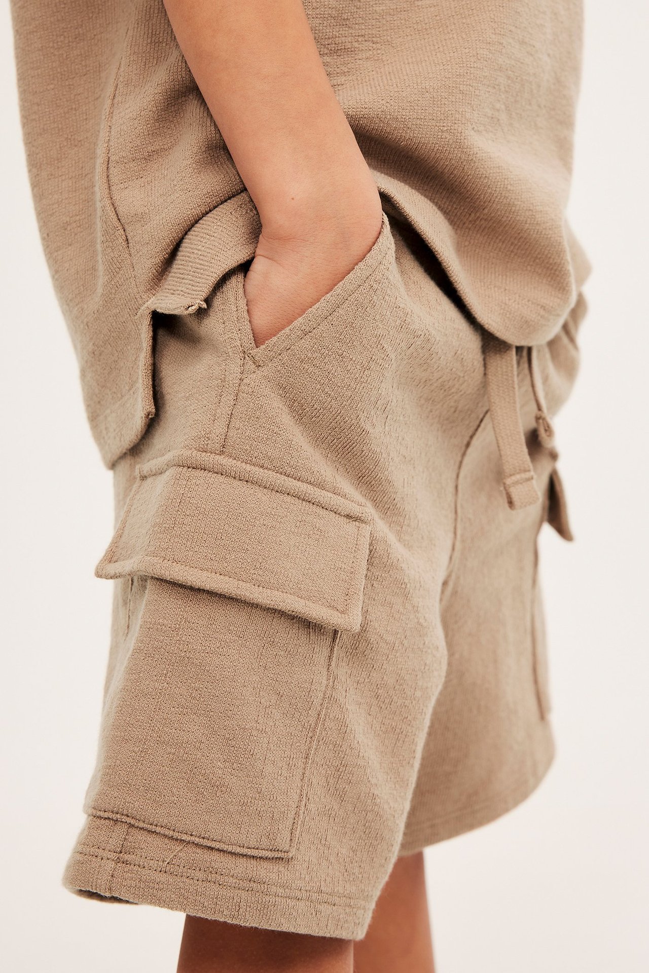 Närbild, sidovy: Barn bär bruna creppade cargoshorts i bomull med handen i fickan. Matchande topp synlig.