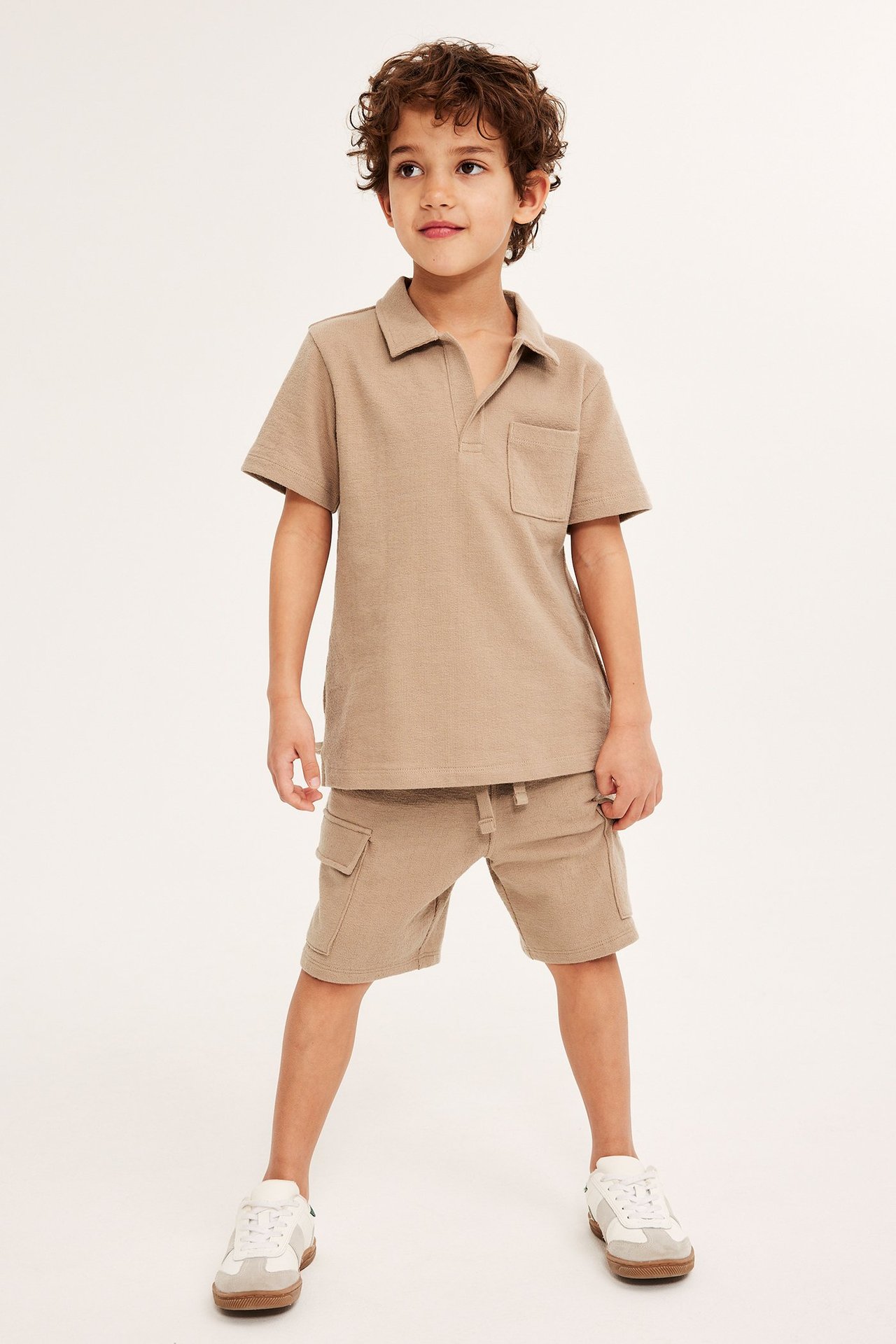 Framifrån, ett barn i ljusbruna cargoshorts i bomull med fickor, matchande pikétröja och vita sneakers.