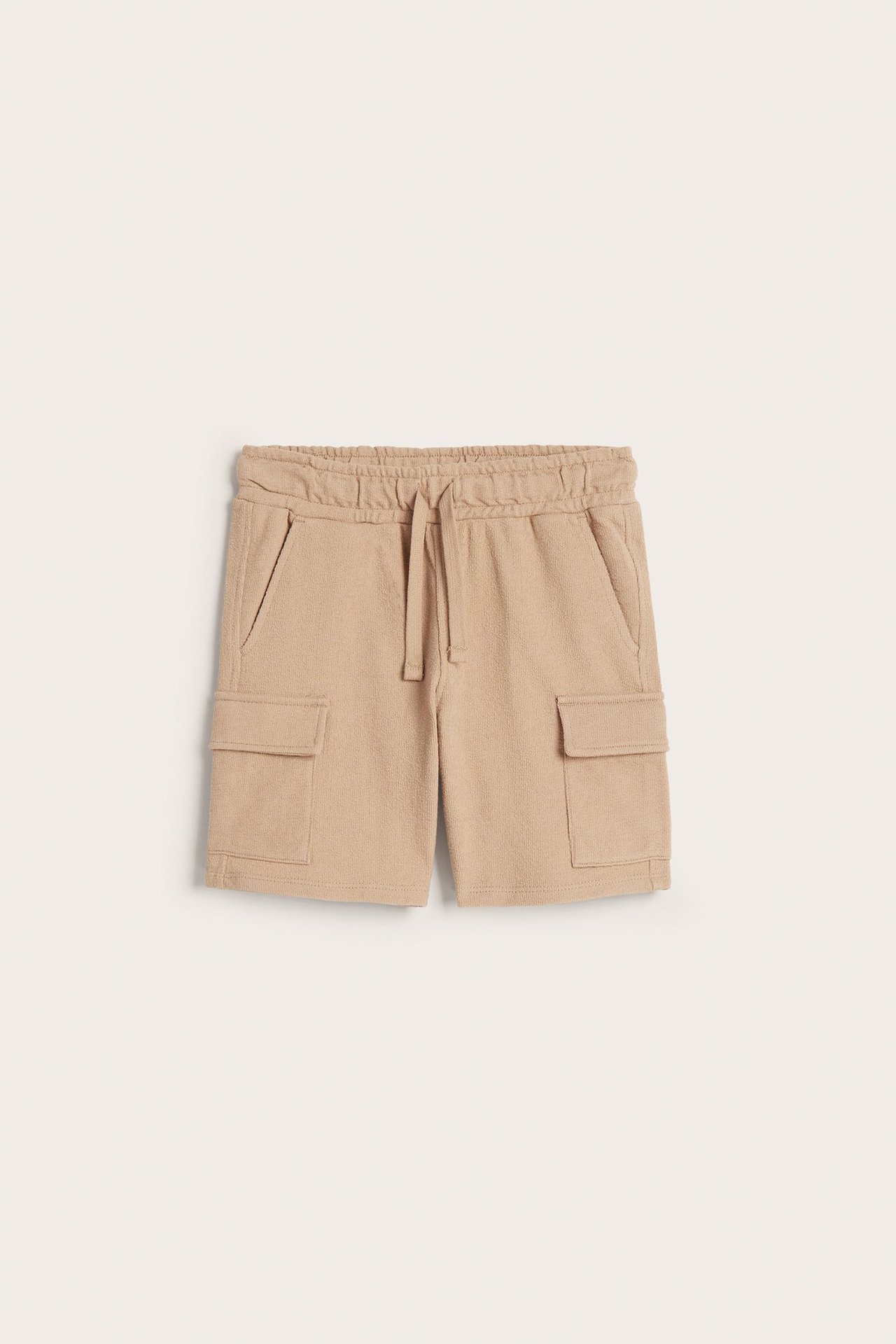 Framifrån: beiga cargoshorts i bomull för barn med resår i midjan, dragsko och fickor med lock.