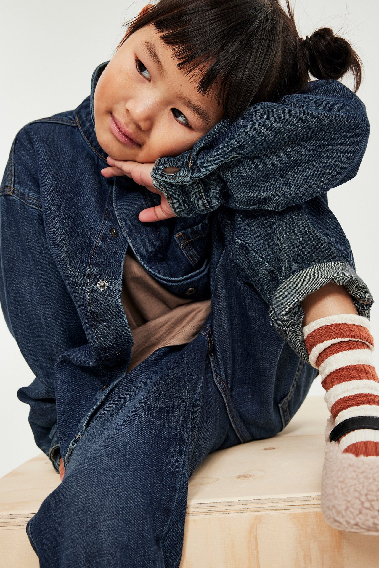Barn i mörk denimblå jeansskjorta öppen över brun t-shirt, jeans, randiga strumpor, toffel.