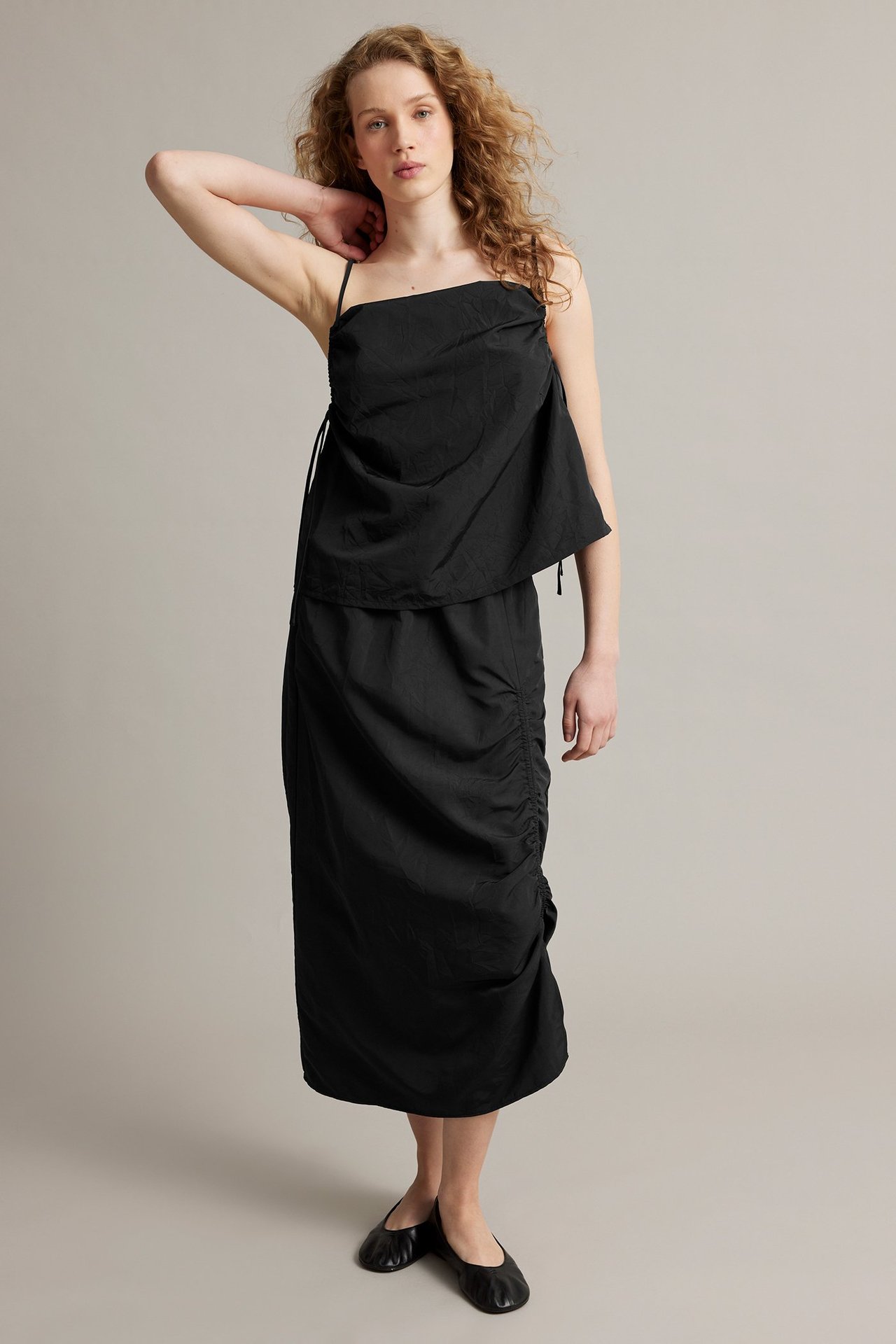 Frontvy av kvinna i svart vävt draperat linne, matchande draperad kjol och svarta ballerinaskor.