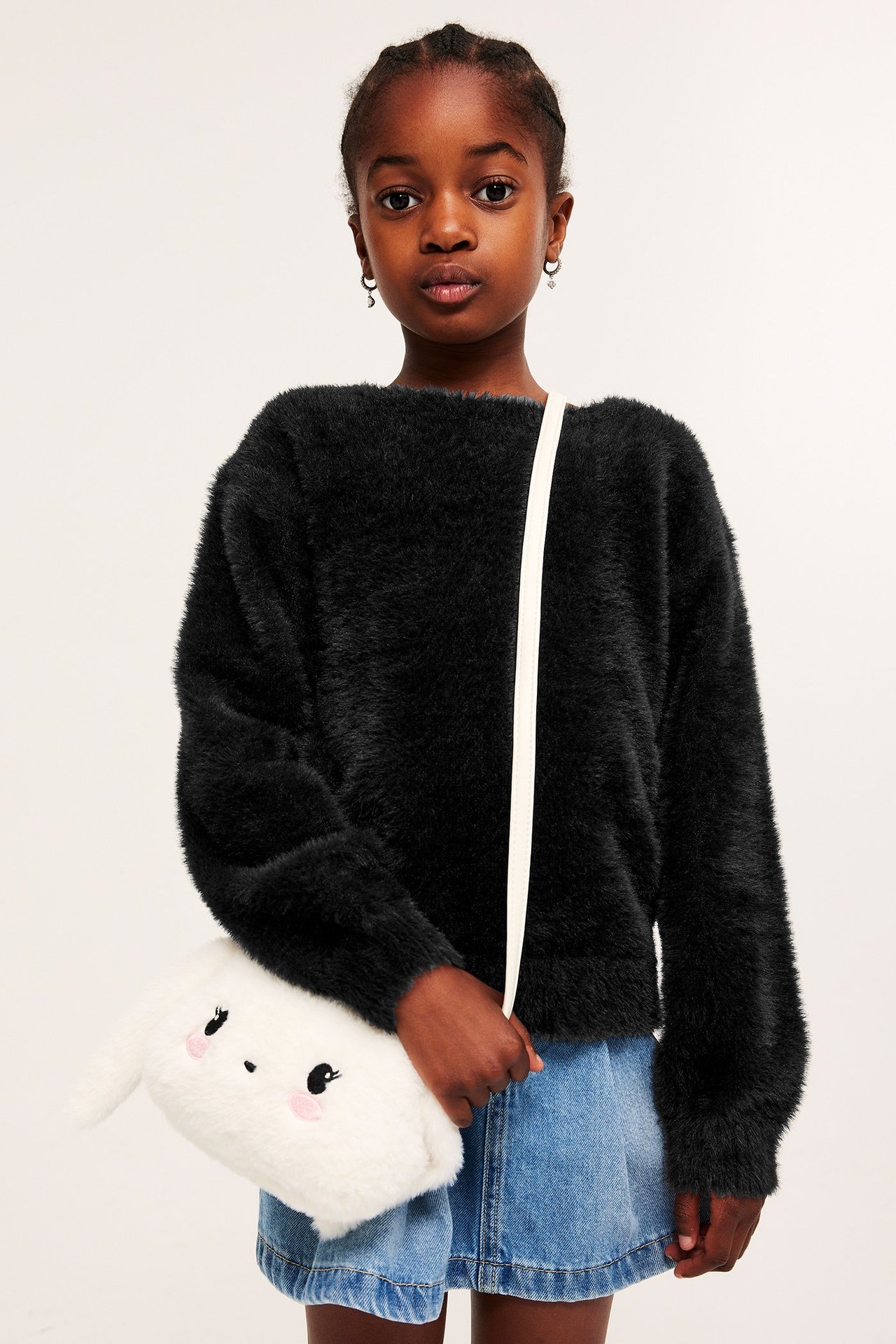 Forfra: Barn i svart strikket genser i feather yarn, blått dongeriskjørt, holder hvit kaninveske.