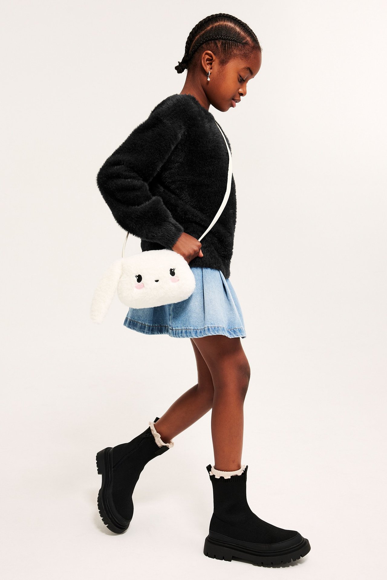 Widok z boku dziecka w czarnym swetrze z przędzy feather yarn, jasnoniebieskiej spódnicy jeansowej, czarnych butach i białej torbie króliku.