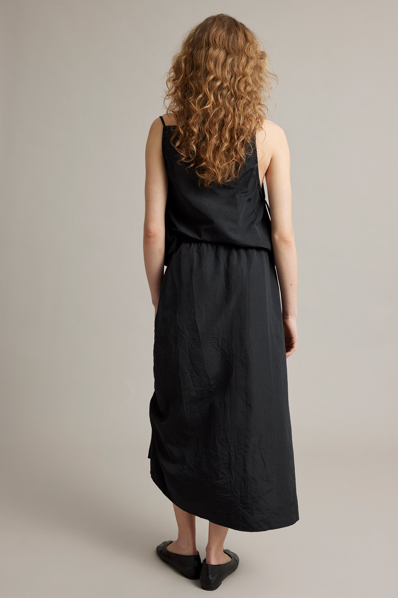 Ryggvy: kvinna i svart draperad midikjol, svart ärmlöst linne och svarta platta skor.