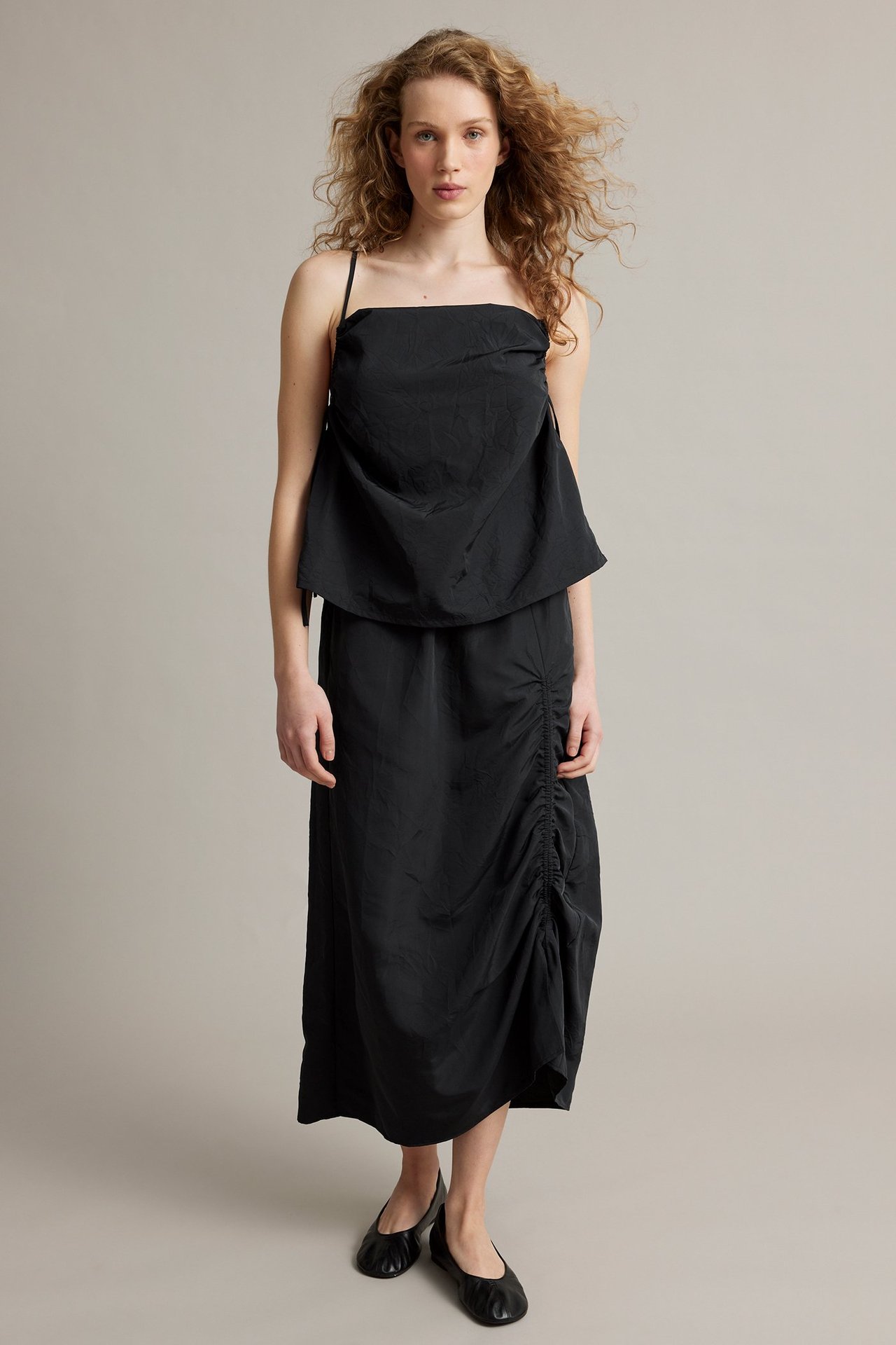 Framifrån, kvinna i svart midikjol med justerbar sidodrapering, svart camisole och svarta platta skor.