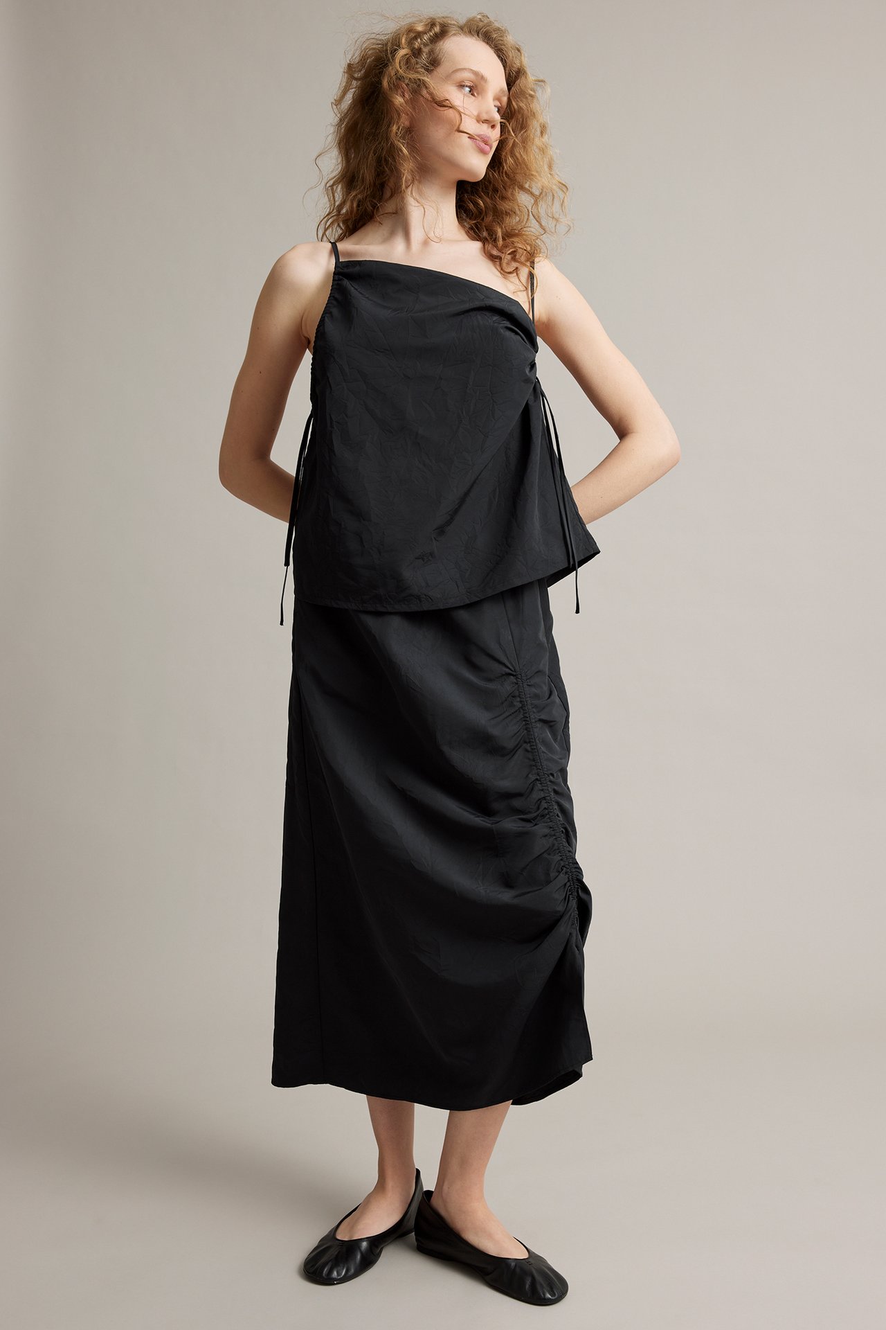 Widok z przodu kobiety w czarnej spódnicy midi z bocznymi marszczeniami, czarnym topie i czarnych balerinach.