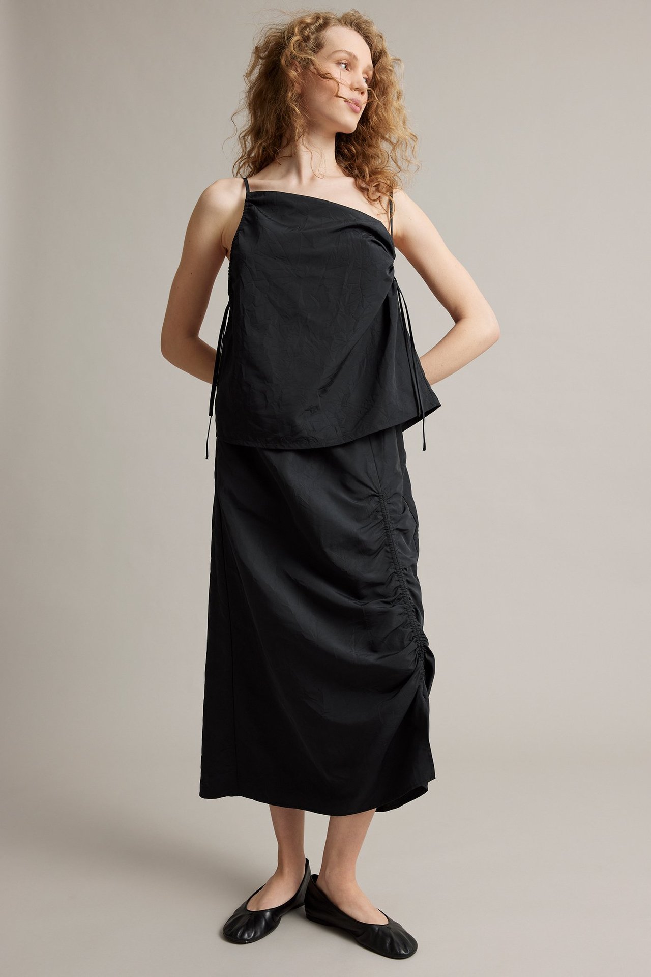 Widok z przodu kobiety w czarnej spódnicy midi z bocznymi marszczeniami, czarnym topie i czarnych balerinach.