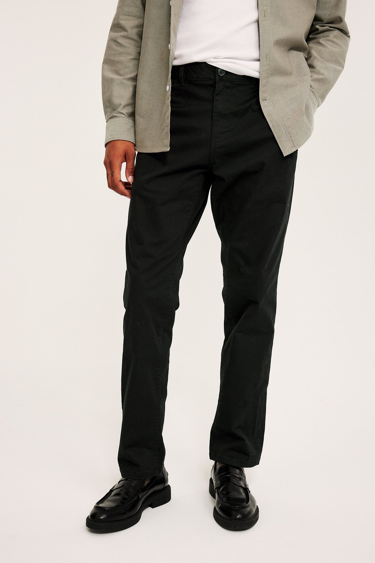 Framifrån: man i svarta regular fit chinos, vit t-shirt, ljusgrå skjorta och svarta loafers.