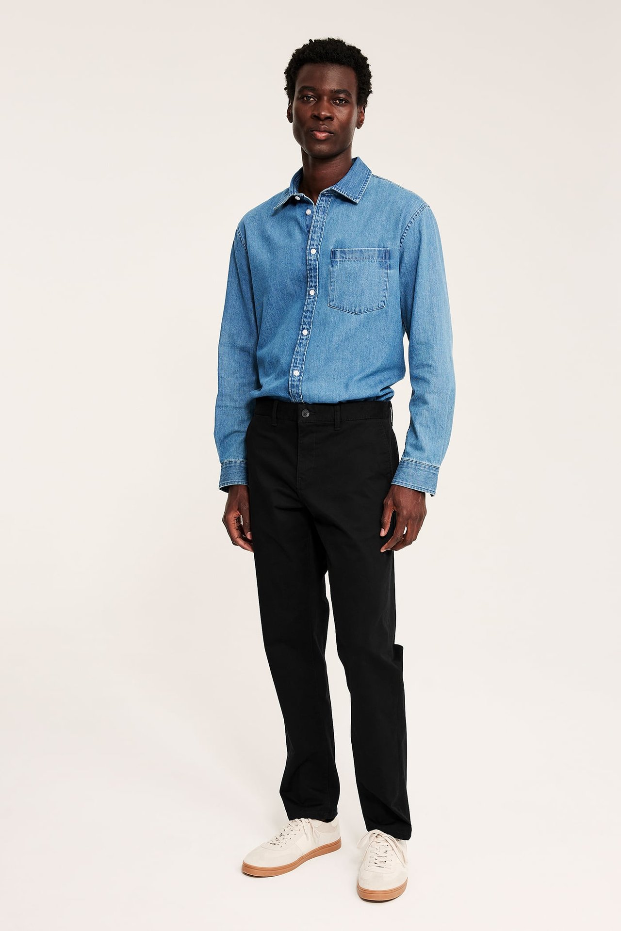 Framifrån, manlig modell i blå långärmad denimskjorta, svarta byxor och beiga sneakers.