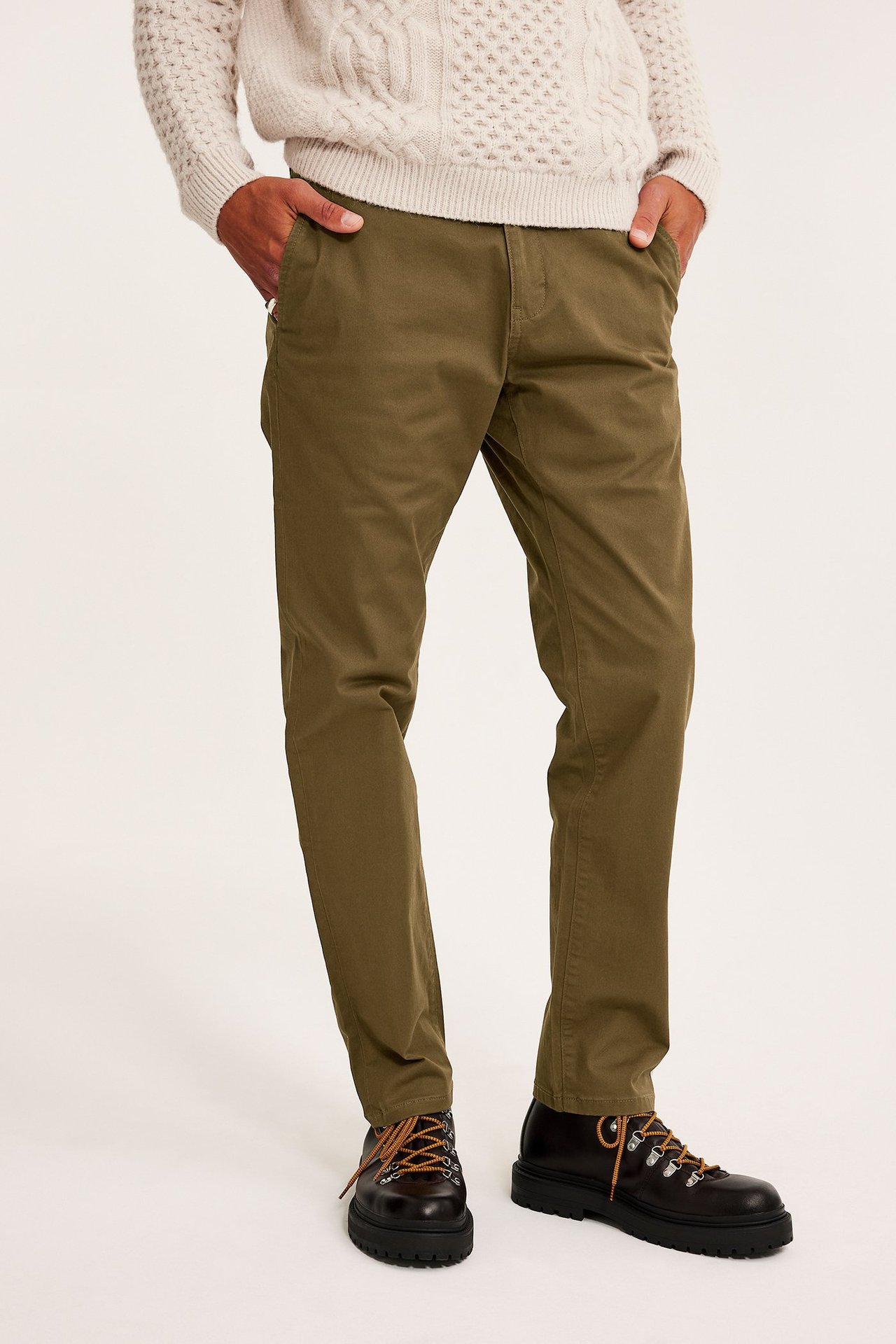 Frontvisning av en mann i brune regular fit chinos, en kremhvit flettet genser og mørkebrune støvler.