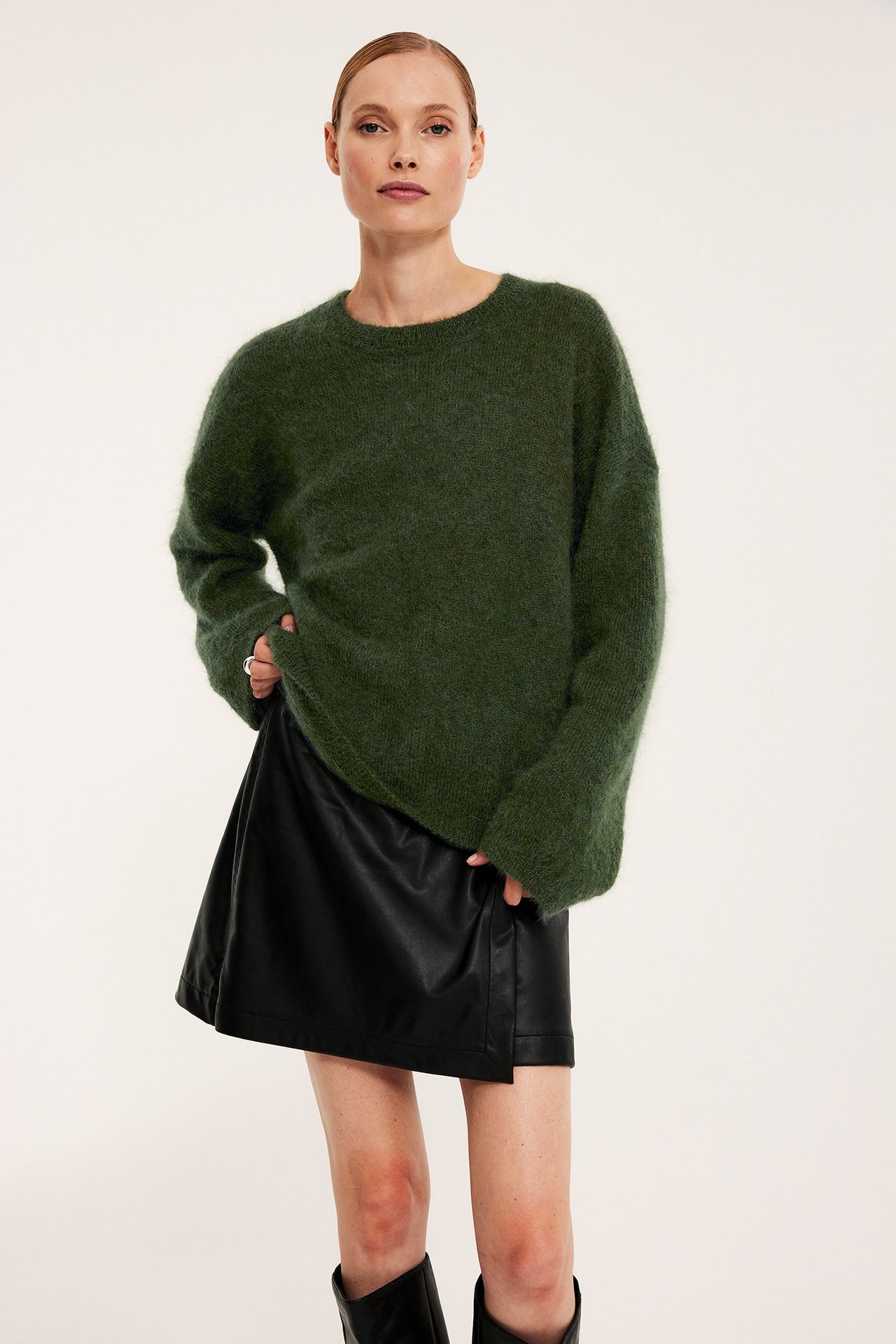 Edestäpäin: nainen vihreässä oversized mohairsekoite-neuleessa, mustassa tekonahkahameessa ja mustissa saappaissa.