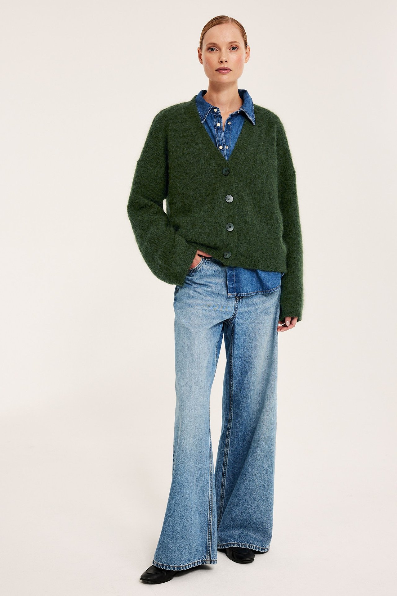 Kobieta w zielonym kardiganie, jeansowej koszuli, jasnych szerokich jeansach, czarnych butach.