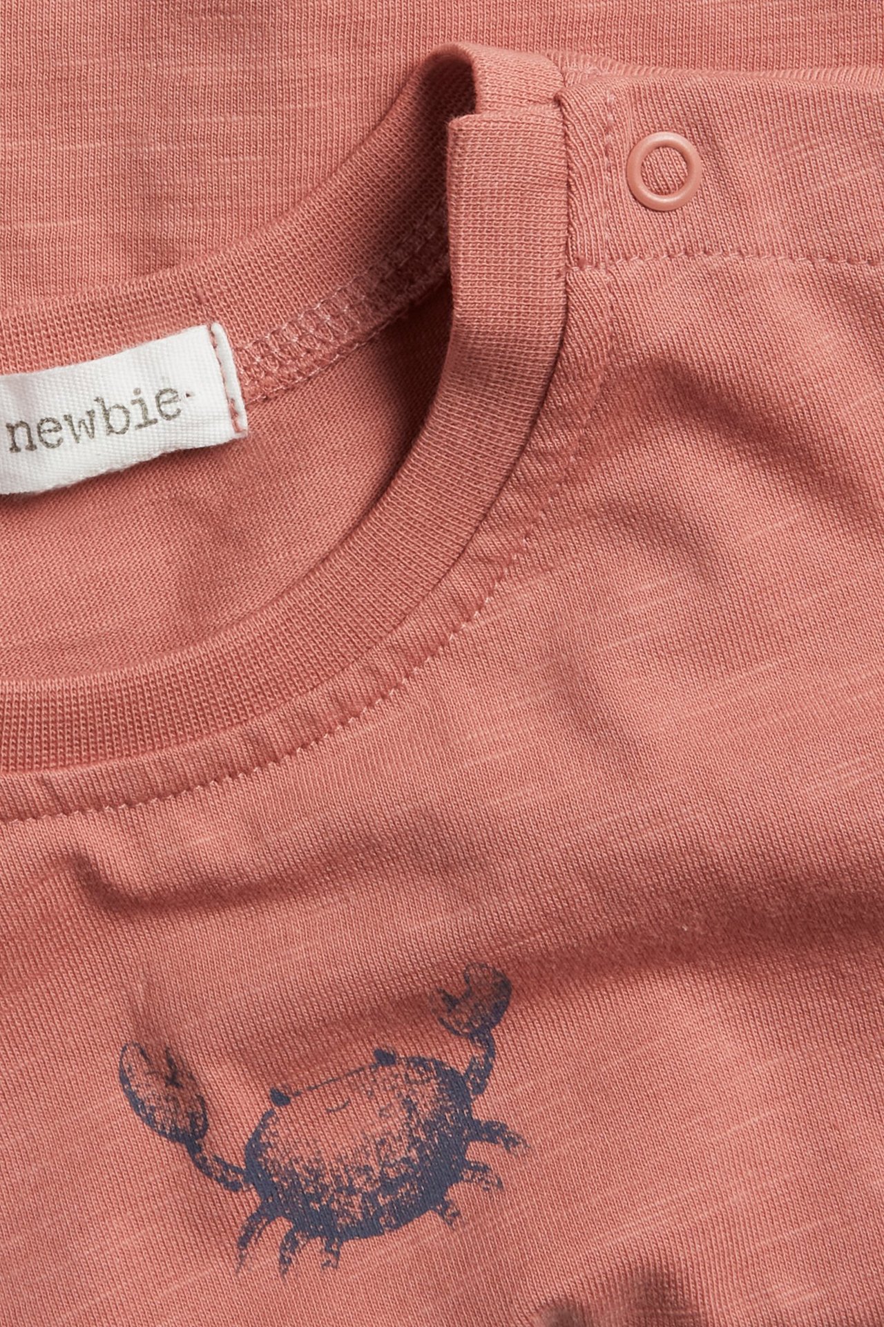 Nærbilde av en rosa bomulls slub t-skjorte med mørkt krabbetrykk og trykknapp på skulderen.