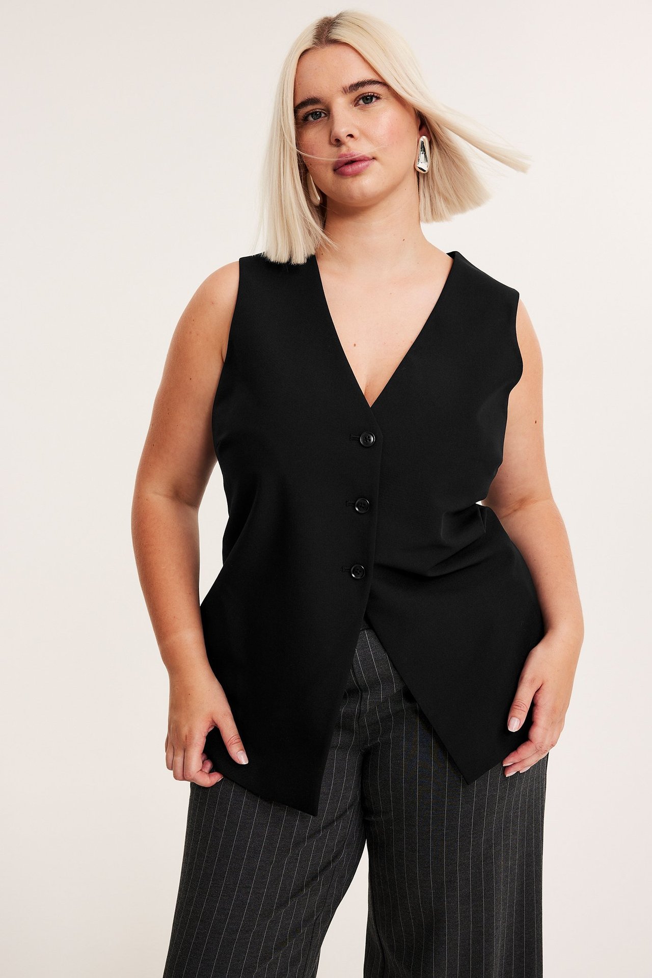 Edestä näkymä naisesta mustassa V-aukkoisessa napitetussa liivissä, tummanharmaissa liituraitahousuissa ja hopeakorvakoruissa.