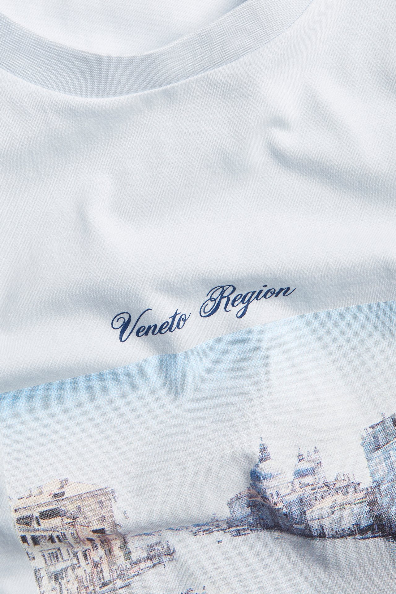 Närbild framifrån av en ljusblå bomulls-t-shirt med Venedig-stadsmotiv och texten 'Veneto Region'.