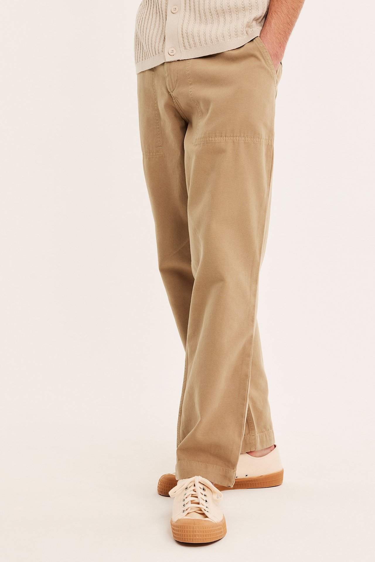 Edestä: Mies beigenvärisissä puuvillaisissa työhousuissa, vaaleanbeigessä ribbineuletakissa ja lenkkareissa.