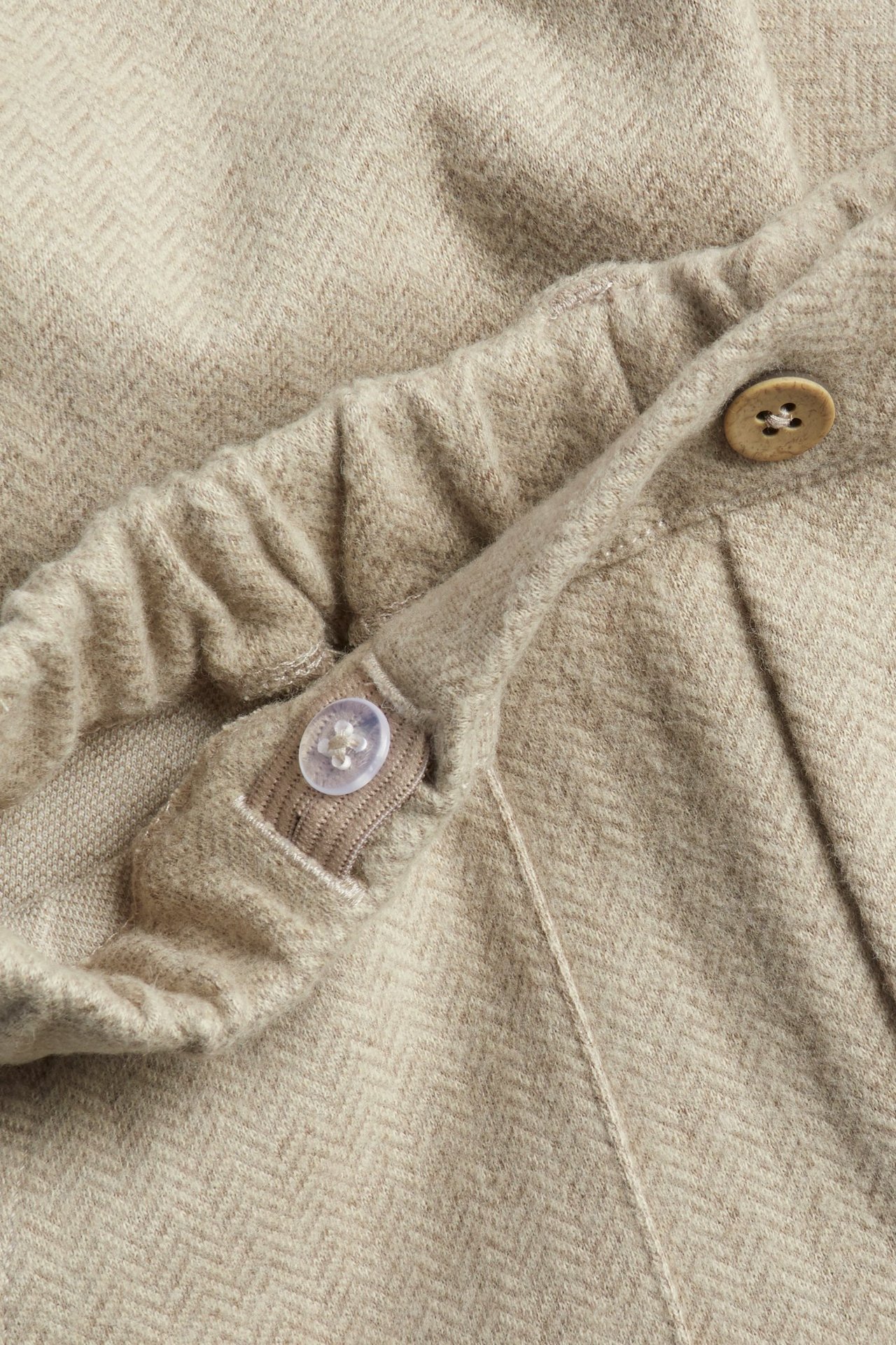 Zbliżenie beżowych melanżowych spodni dziecięcych w jodełkę, z elastyczną talią i ozdobnymi guzikami.