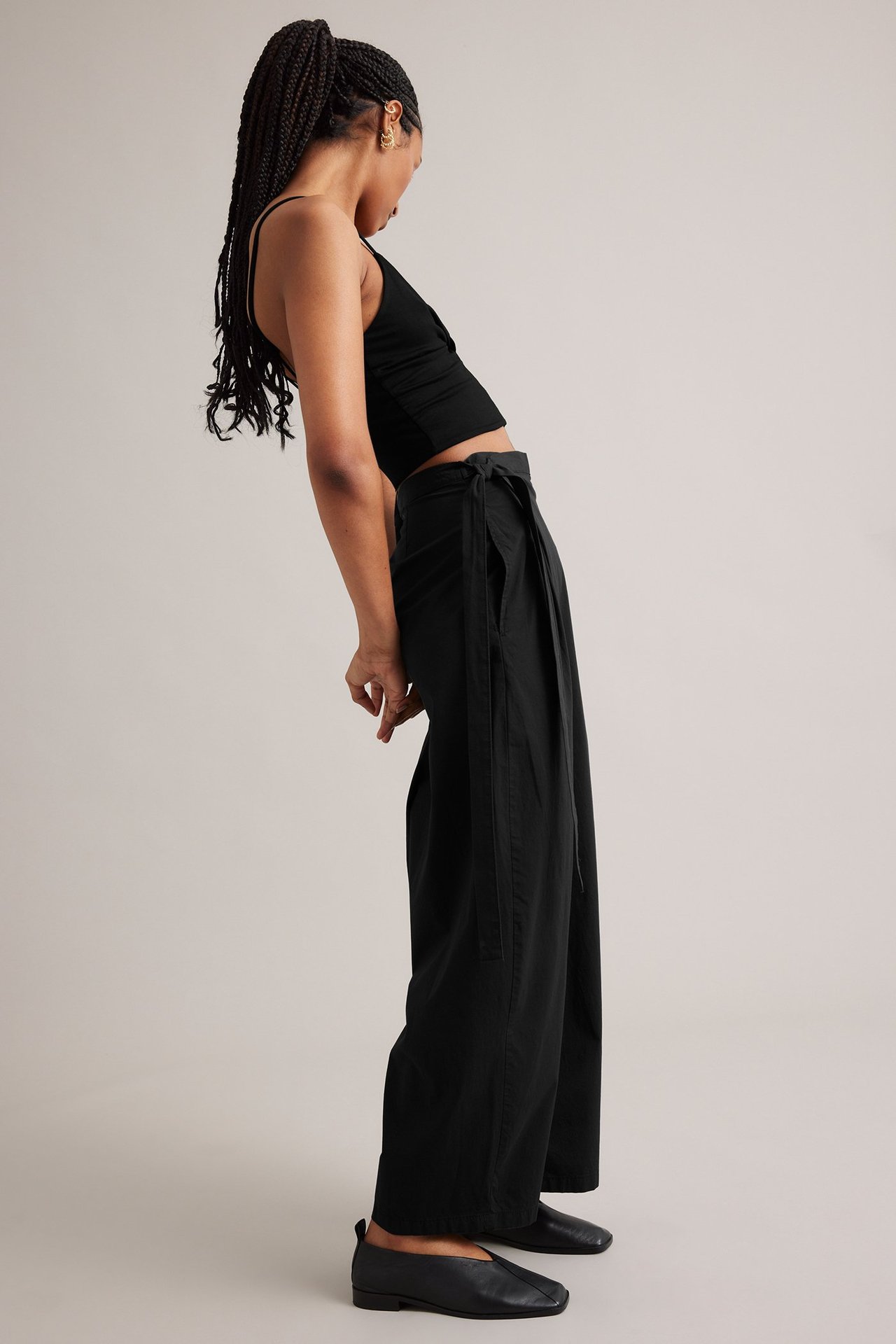 Naisen sivukuva mustissa leveälahkeisissa kietaisuhousuissa, mustassa crop topissa ja mustissa kengissä.