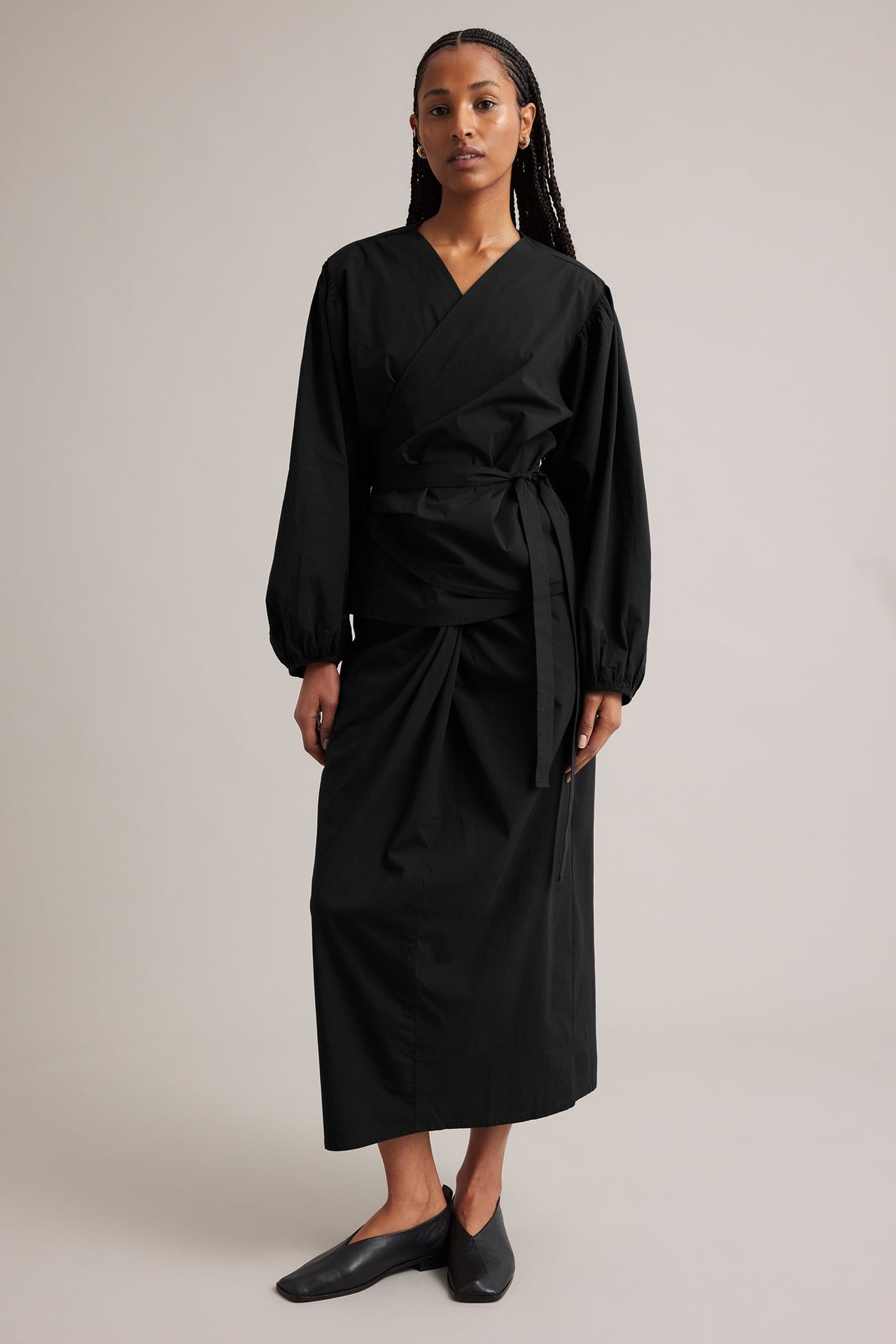 Edestä: Nainen mustassa puuvillasekoite-kietaisublousissa pöyhkeillä hihoilla, mustassa midihameessa ja mustissa kengissä.