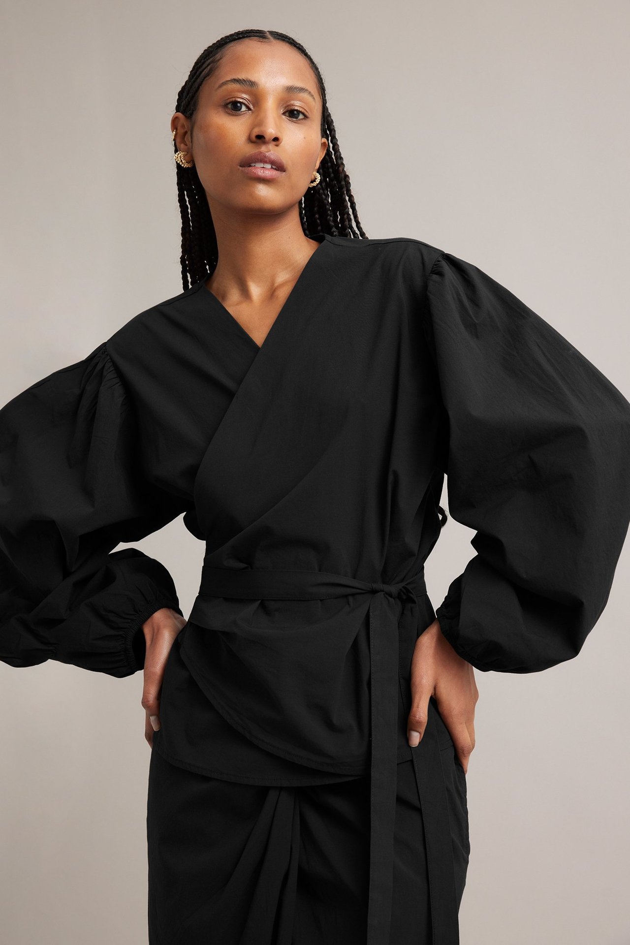 Edestä: nainen mustassa kietaisublousissa, pöyhkeät hihat, sidottu sivuun, mustat housut, kultaiset korvakorut.