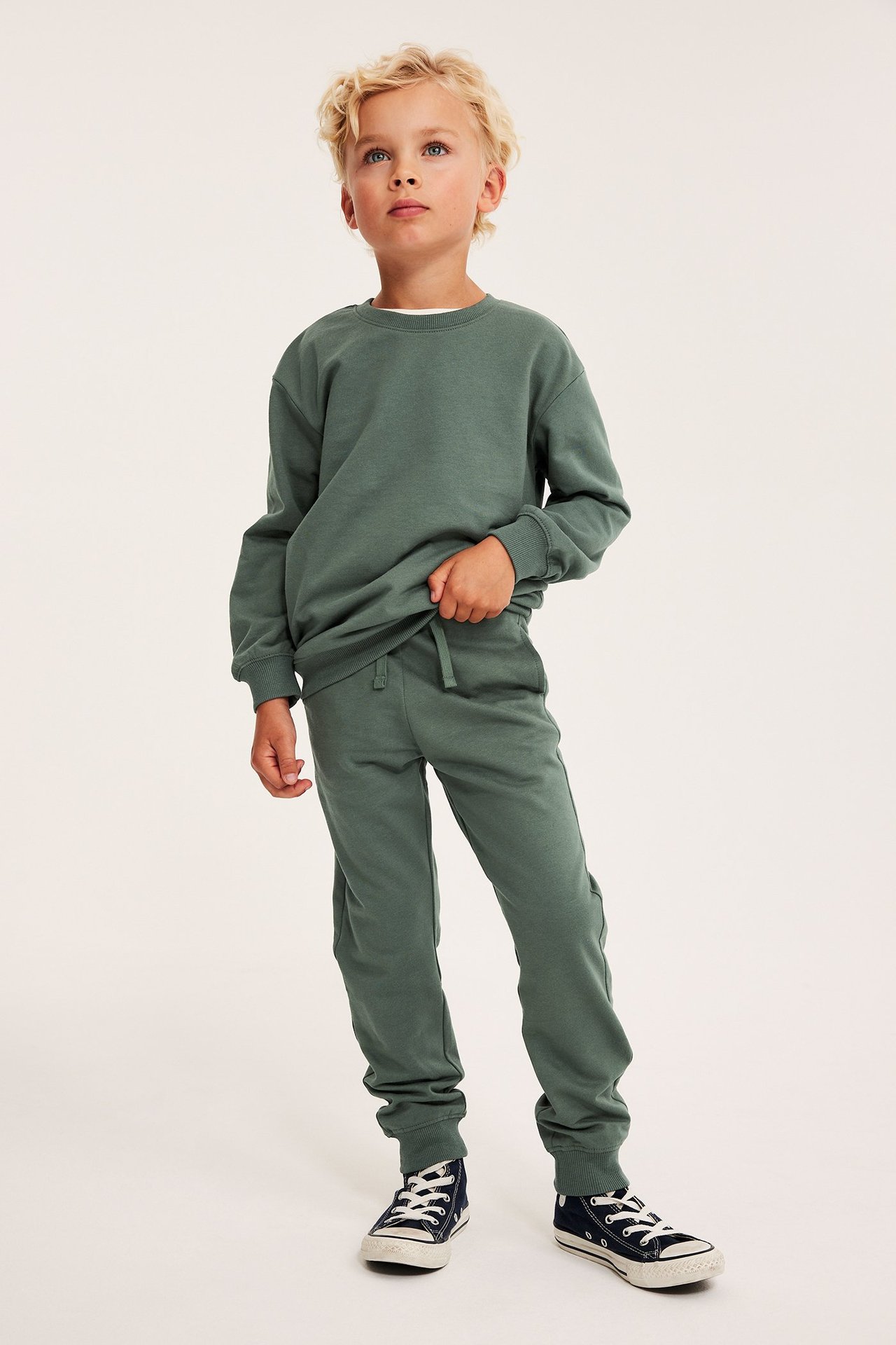 Widok z przodu dziecka w ciemnozielonych bawełnianych spodniach dresowych ze sznurkiem, pasującej bluzie i granatowych trampkach.