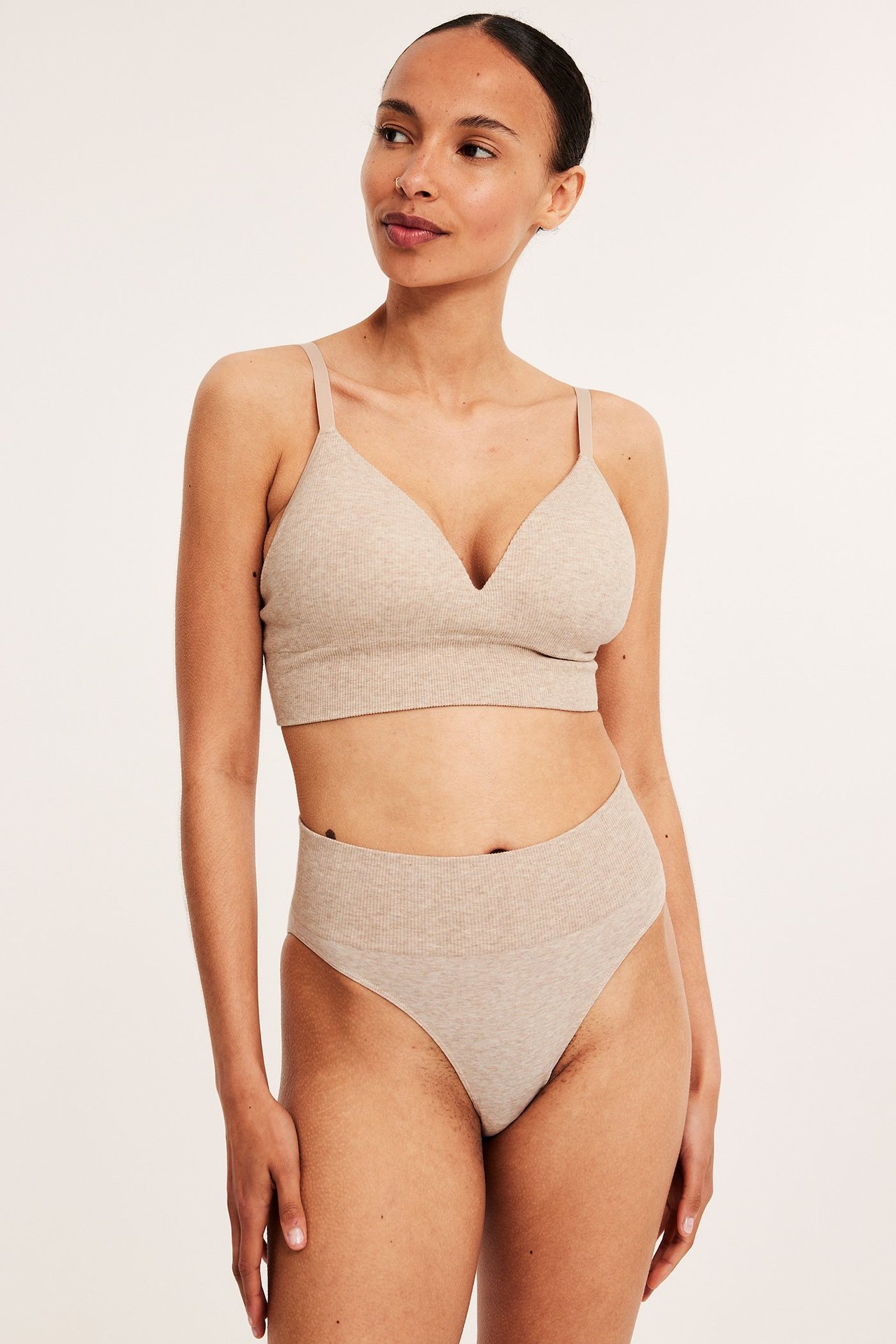 Framifrån: kvinna i beige melerad ribbad sömlös bralette och matchande högmidjade trosor.