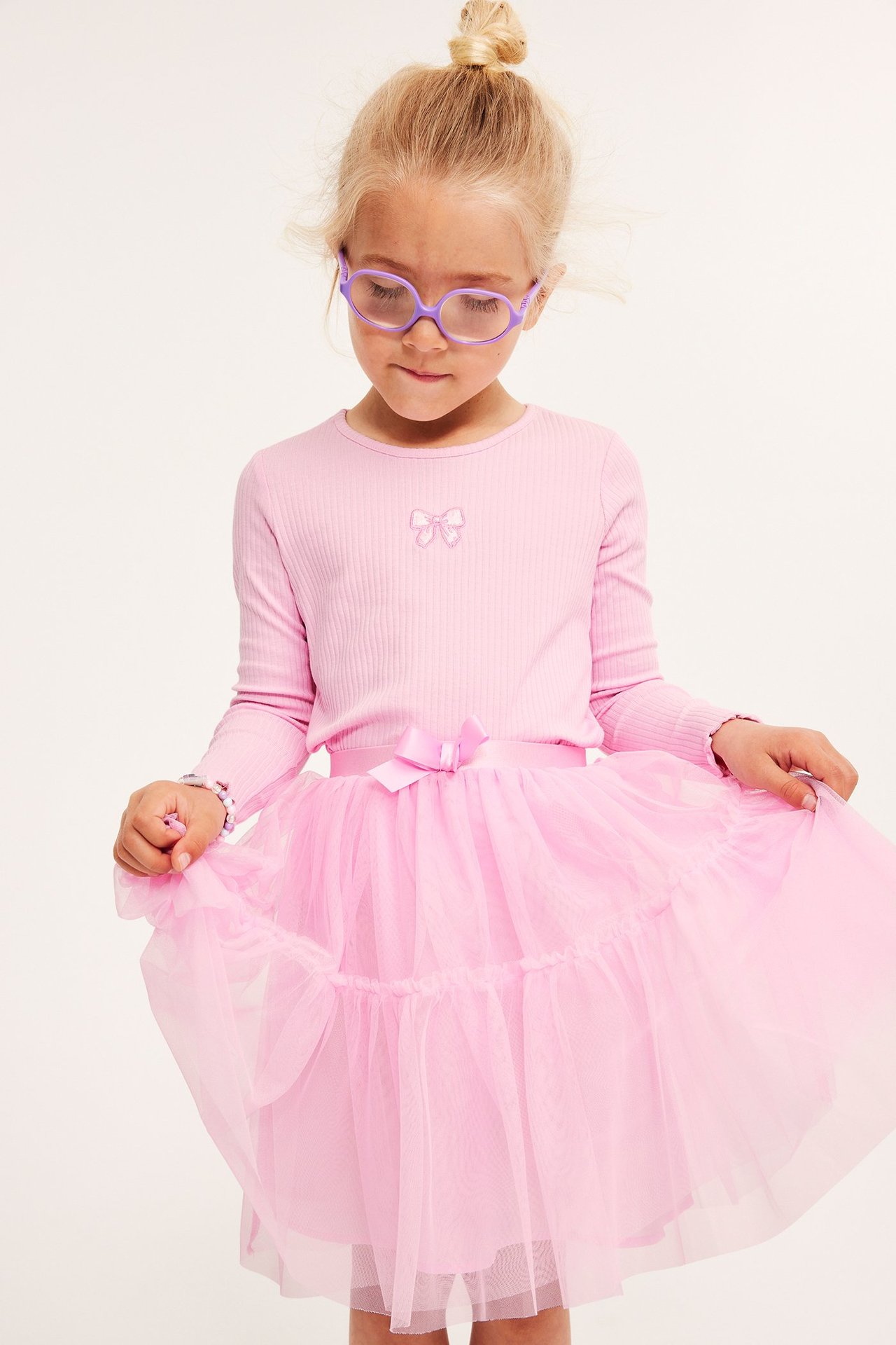 Framifrån: Barn i rosa volangtyllkjol med rosett, rosa ribbad topp, lila glasögon och armband.