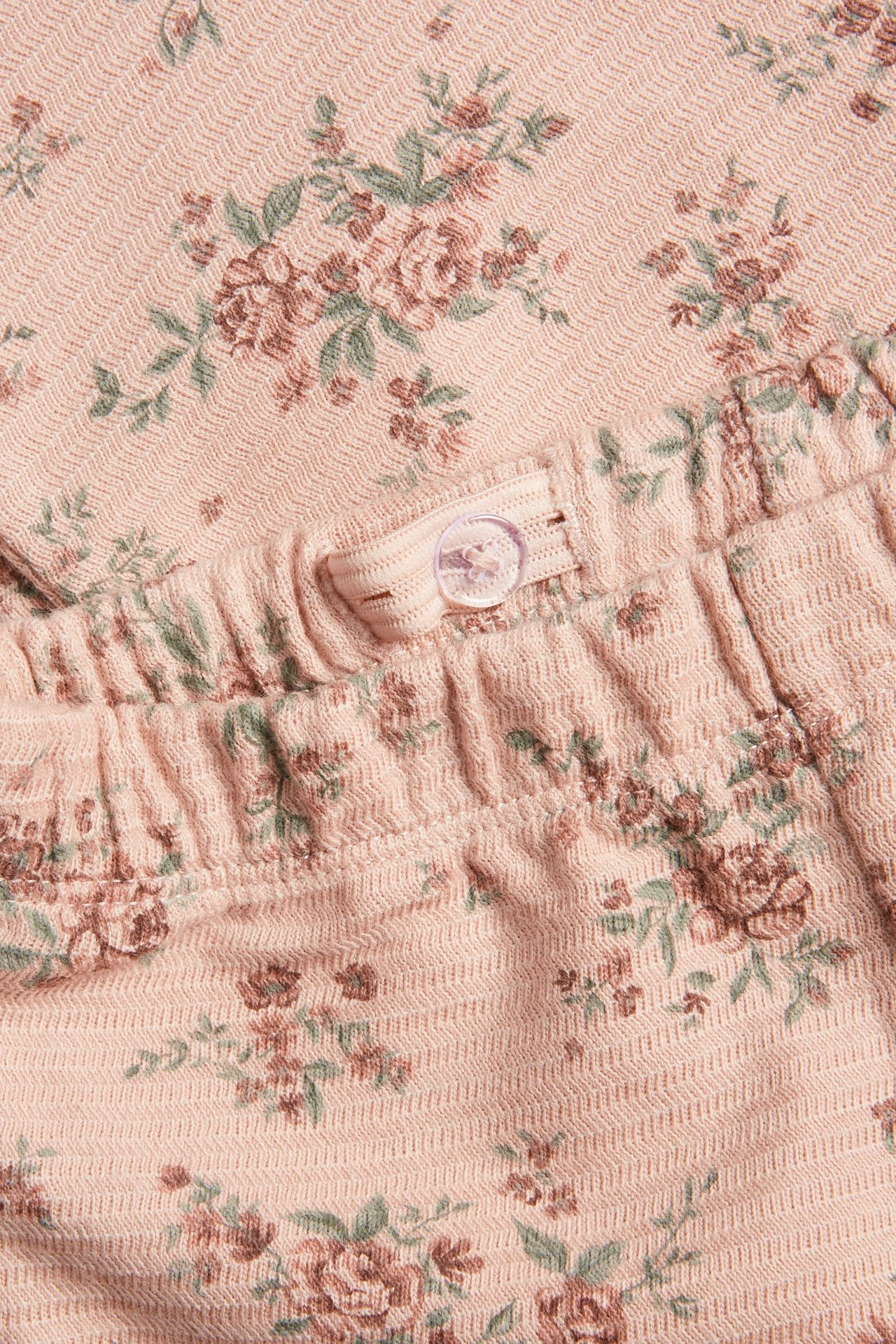 Zbliżenie różowych legginsów z dzianiny strukturalnej w drobny kwiatowy wzór, z regulowaną gumką i guzikiem.