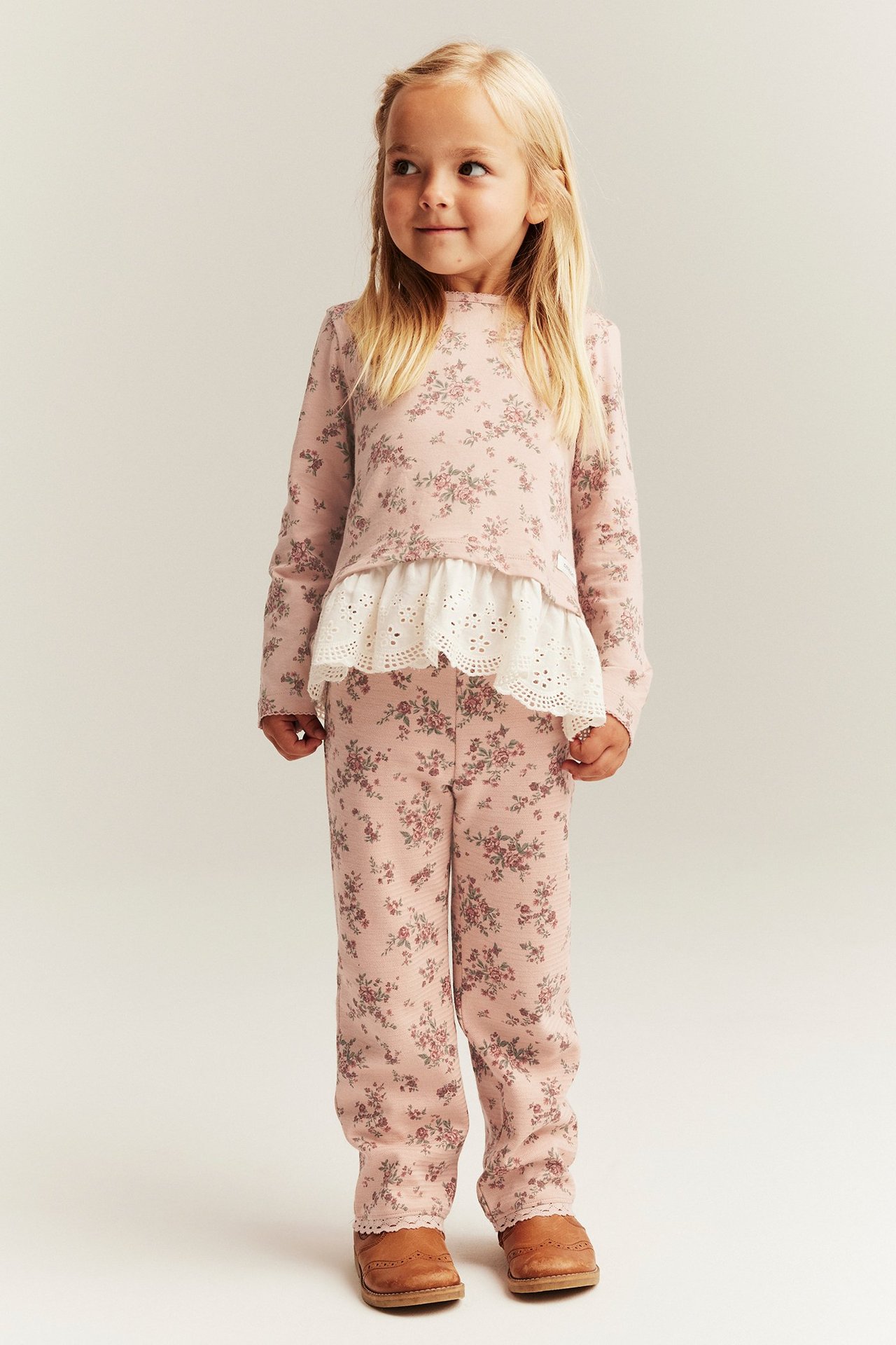 Widok z przodu: Dziecko w różowych kwiecistych rozszerzanych legginsach z koronką, pasującym topie i brązowych butach.