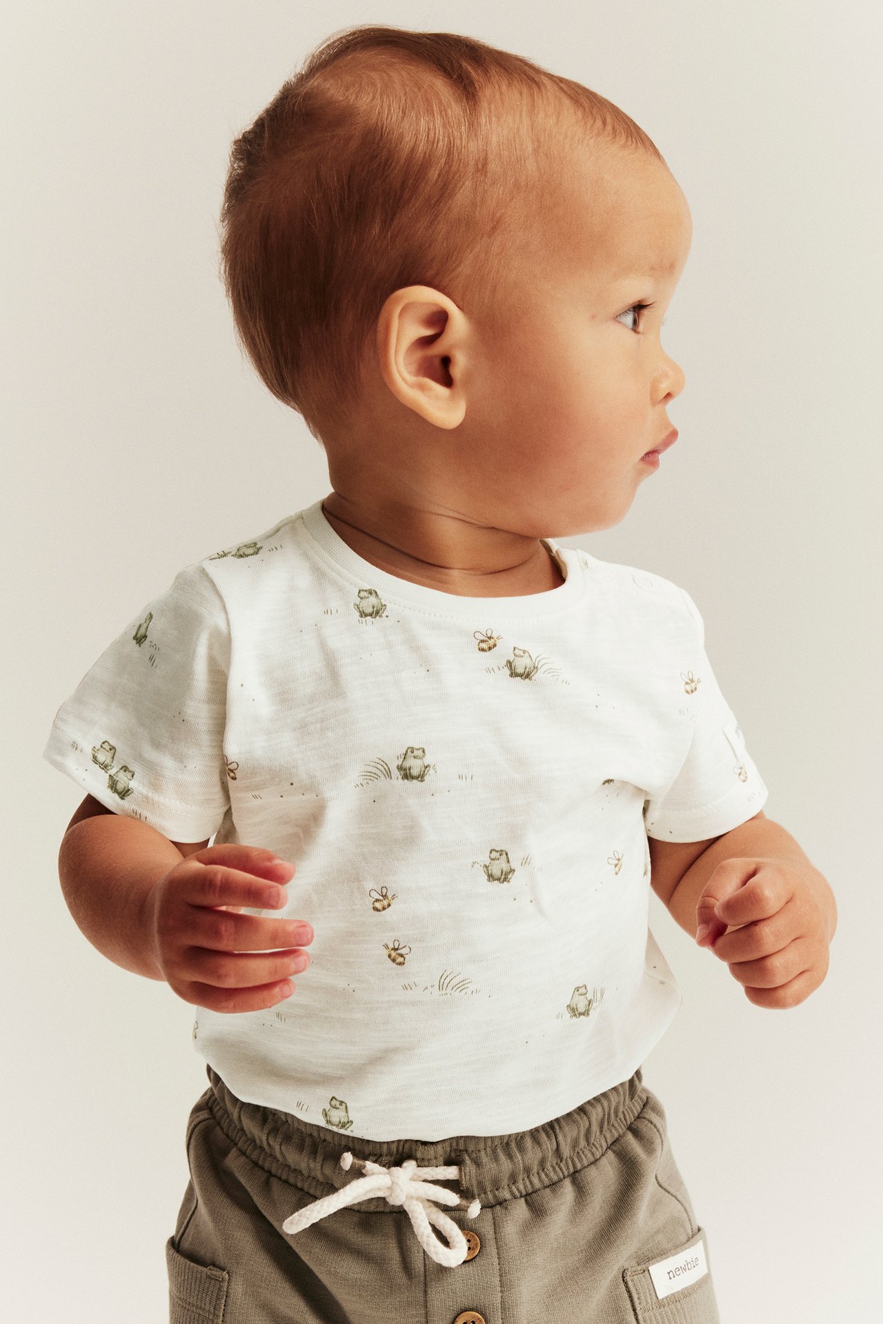 Sidovy av baby i offwhite kortärmad body med grodmönster och grå byxor.