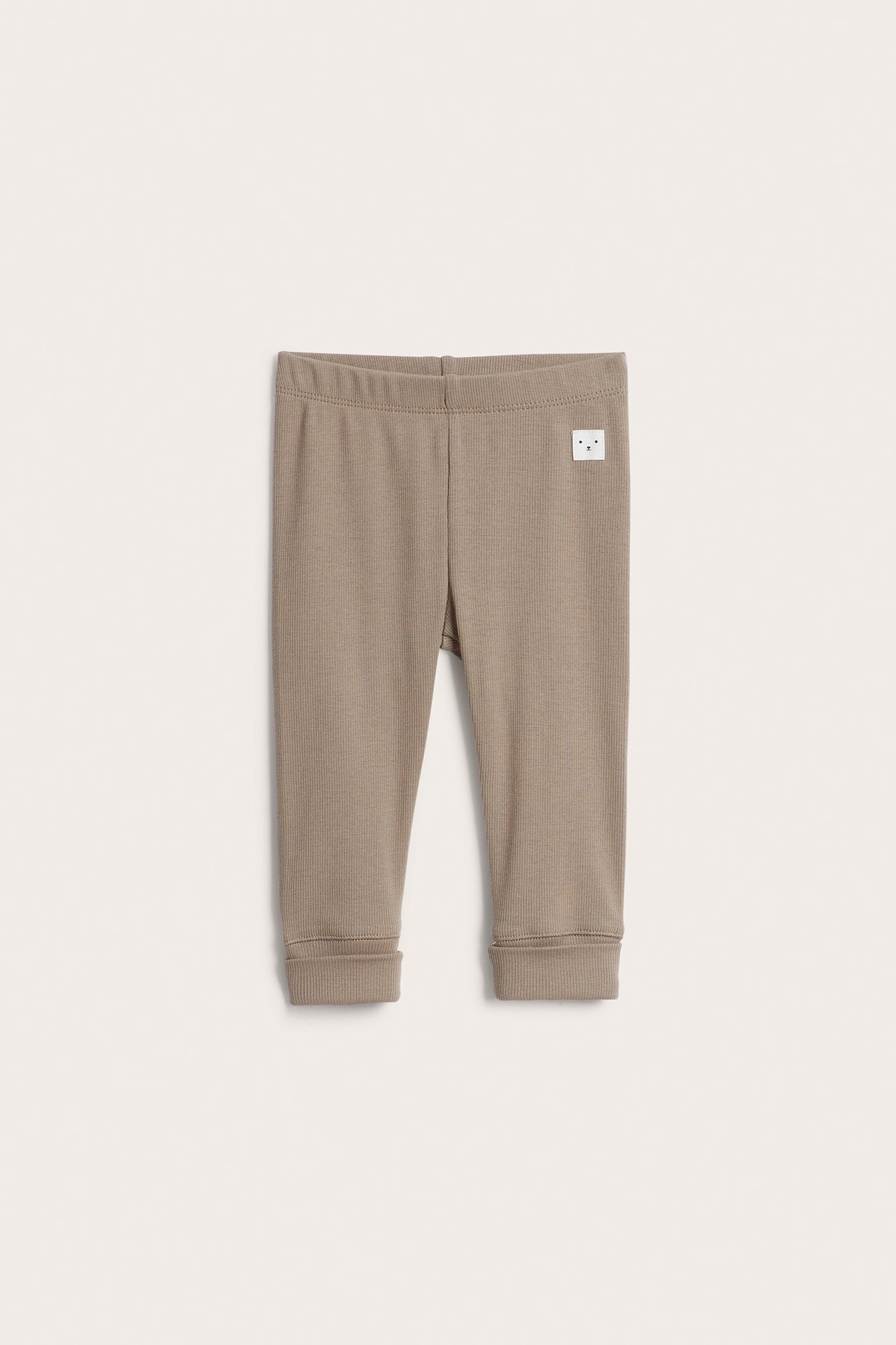 Frontvy av bruna ribbade baby leggings med resår i midjan, muddar och liten vit etikett.