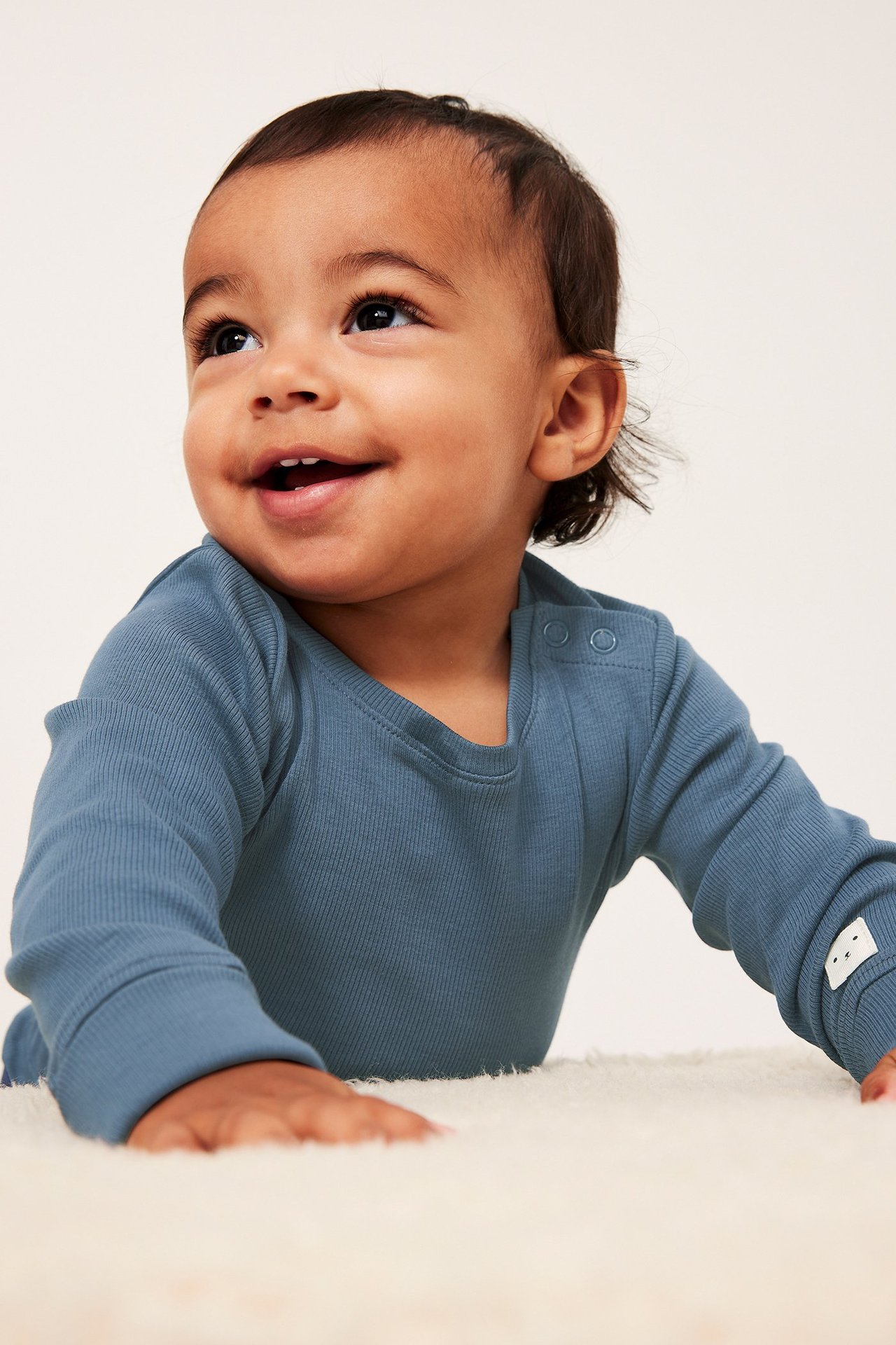 Närbild framifrån på en leende baby i en blå ribbad långärmad body med tryckknappar på axeln.