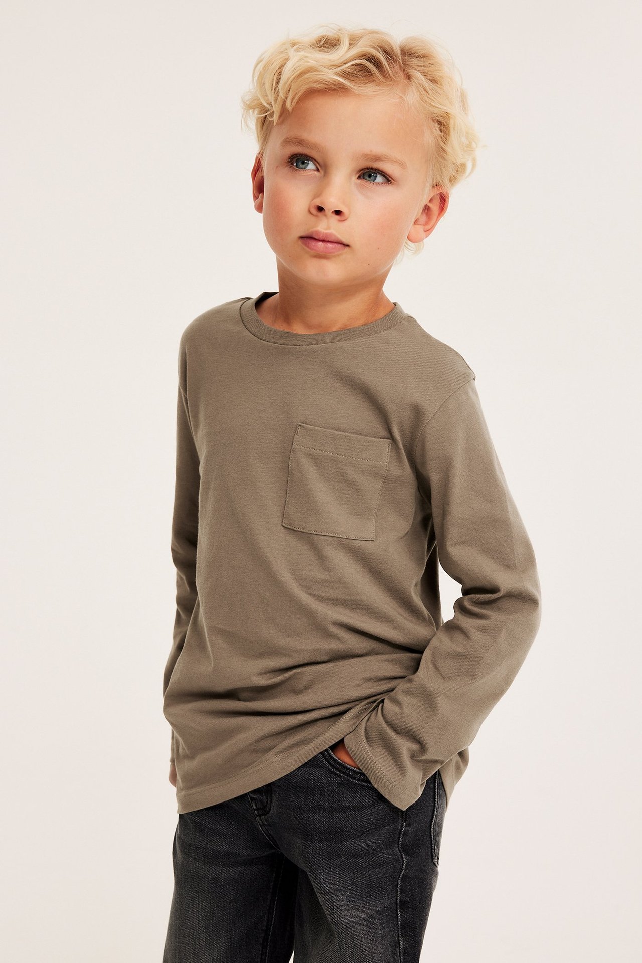 Edestä: Poika khakinruskeassa pitkähihaisessa puuvilla-T-paidassa rintataskulla ja tummanharmaissa farkuissa.